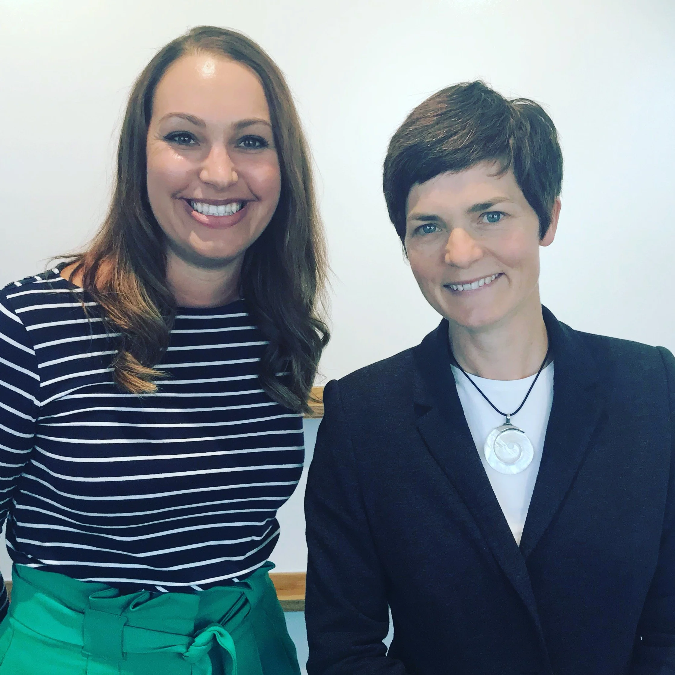Suzy Greenwood and Dame Ellen MacArthur. Two women smiling and standing side by side in front of a white wall, one with long brown hair wearing a striped shirt and green skirt, the other with short dark hair wearing a black blazer and a necklace.