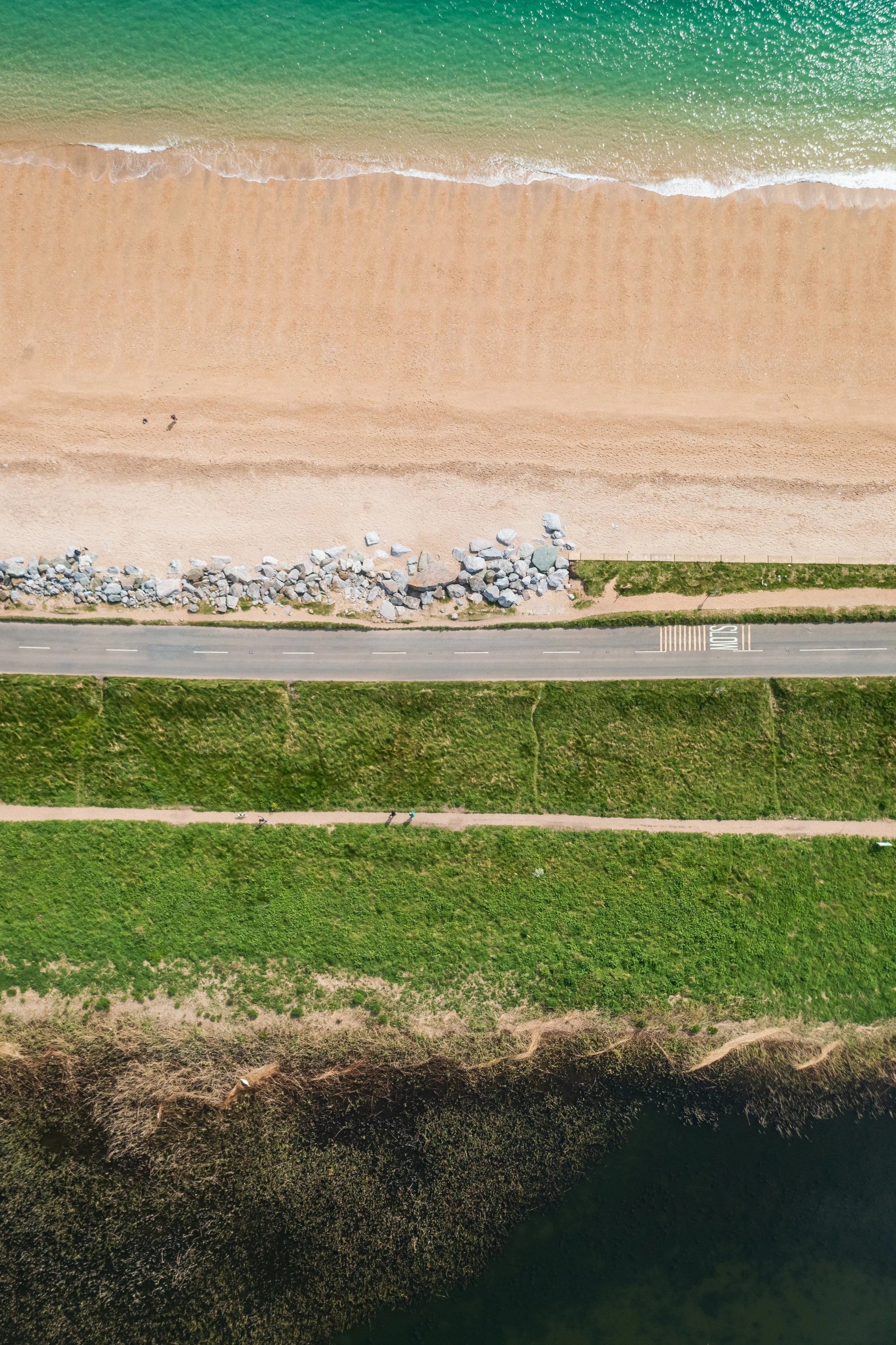 Aerial view of a beach with a sandy shoreline, rocks, a grassy area, a paved road, and water.