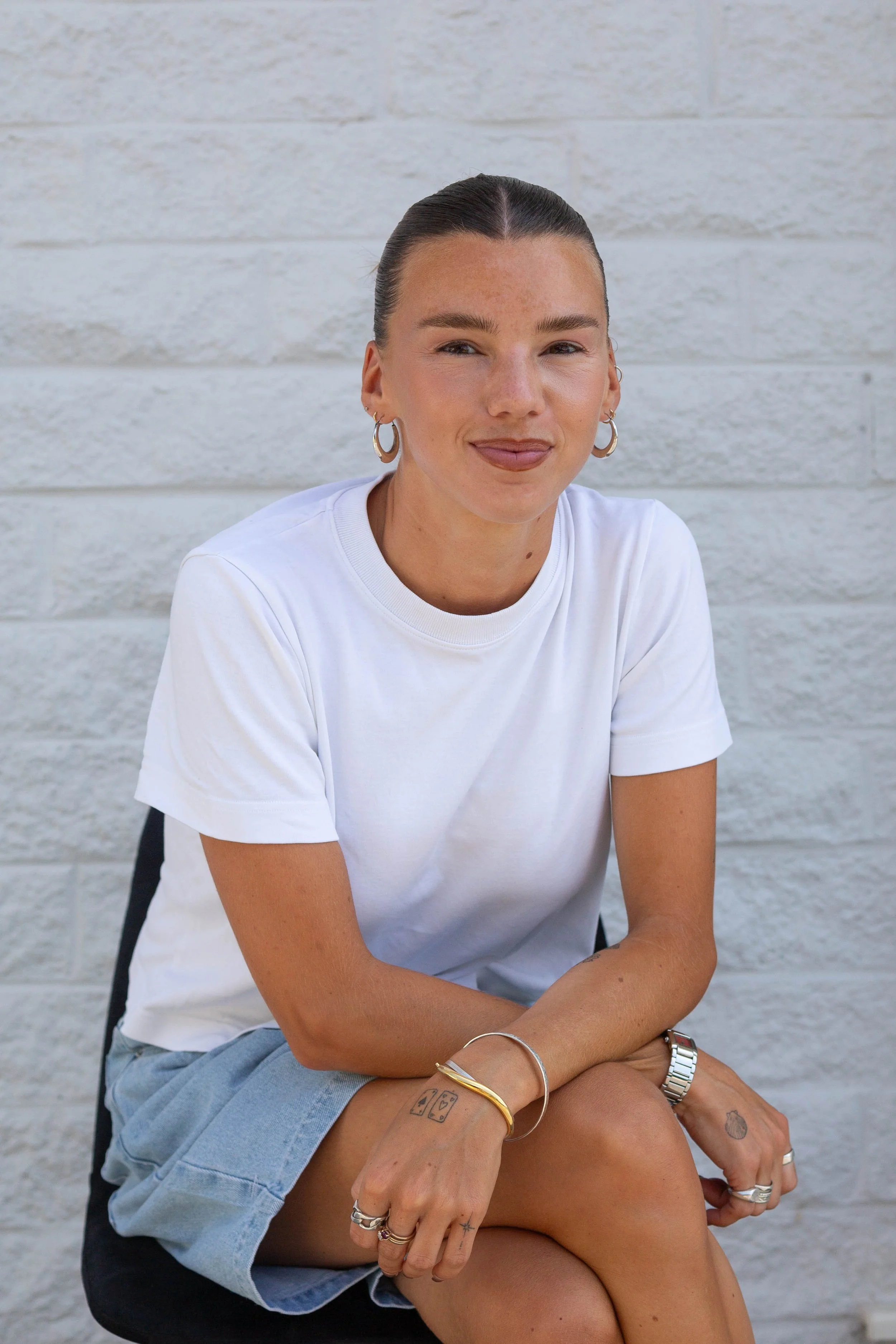 A woman sitting on a person's lap against a white brick wall. She has dark hair pulled back, wears hoop earrings, a white t-shirt, and various jewelry including rings, a bracelet, and a watch.