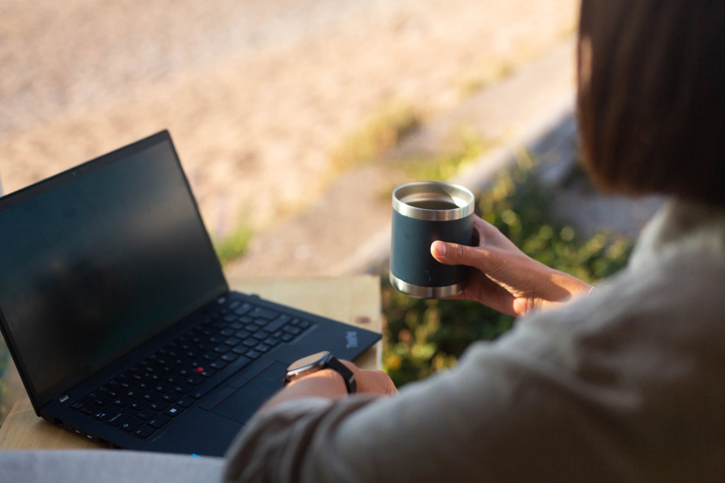 Person holding a coffee mug and working on a laptop outdoors during daylight.