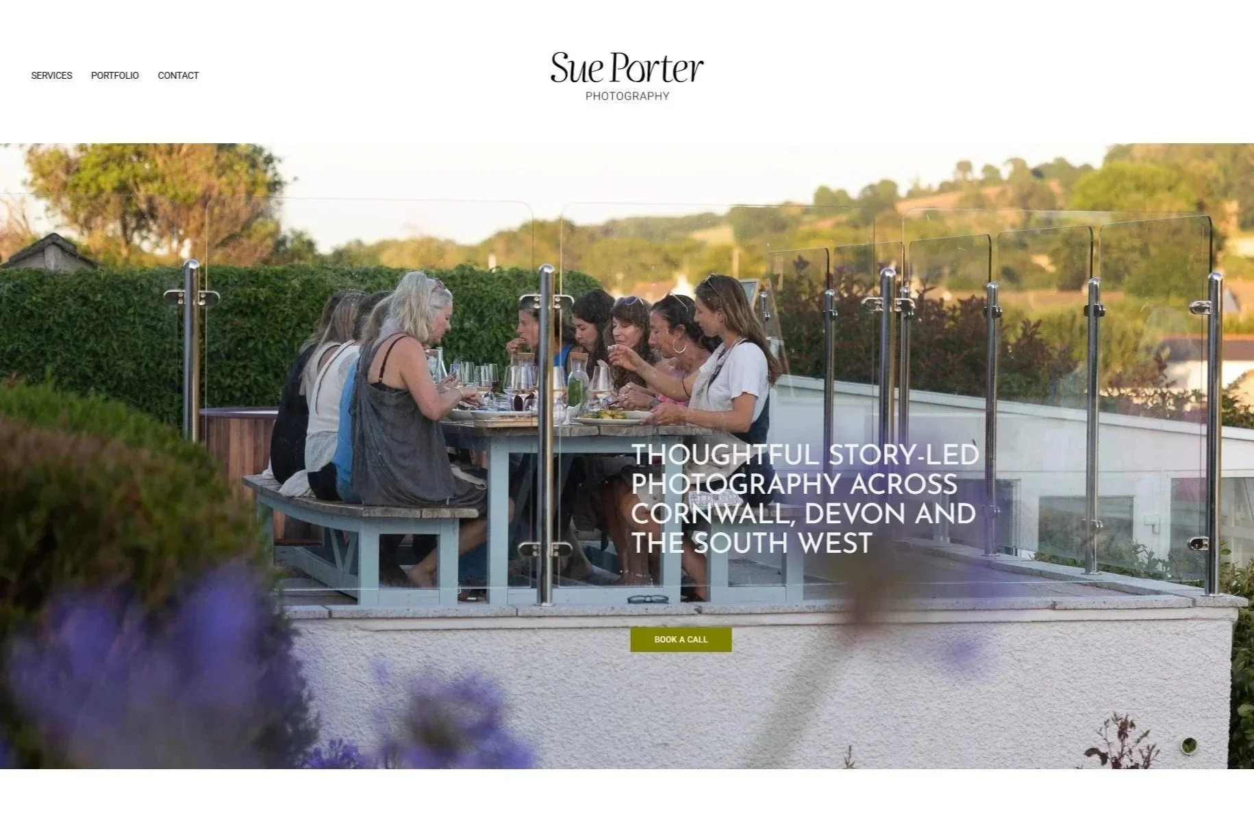 A group of people dining outdoors on a balcony with a glass barrier, overlooking a landscape with rolling hills and trees, during sunset.