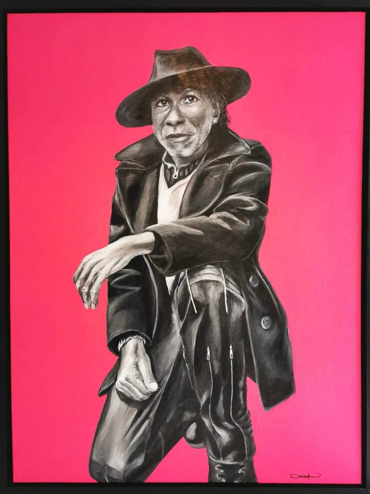 A black and white portrait of a person with a confident expression, wearing a wide-brimmed hat, leather jacket, and pants, posed against a bright pink background.