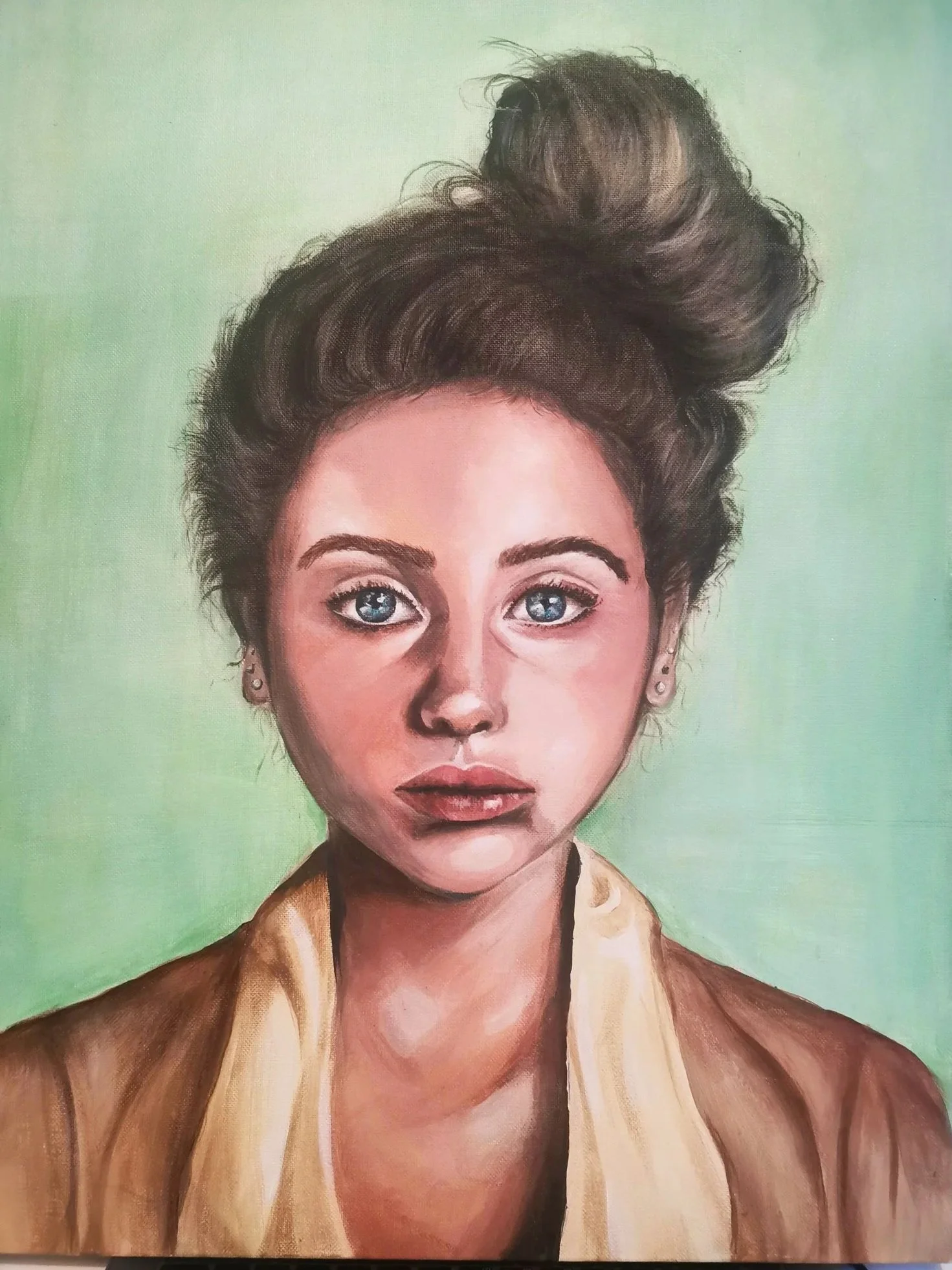 A realistic portrait of a young woman with blue eyes, dark eyebrows, and brown hair styled in a large, messy bun. She is wearing small earrings, a beige shirt, and a brown jacket against a green background.
