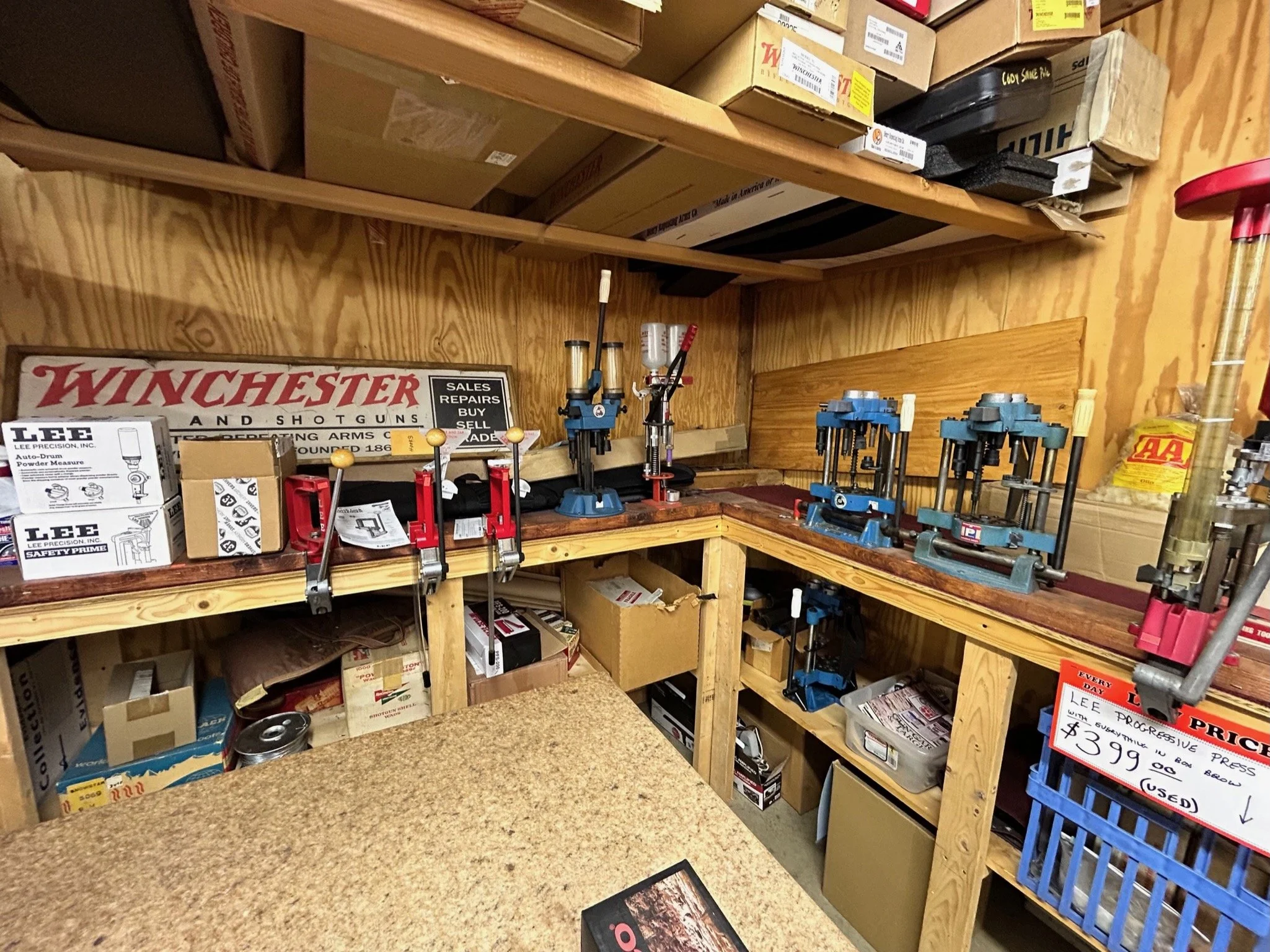 Workbench with reloading presses, boxes, and tools in a gun reloading shop, with Winchester sign and shelves above filled with boxes.