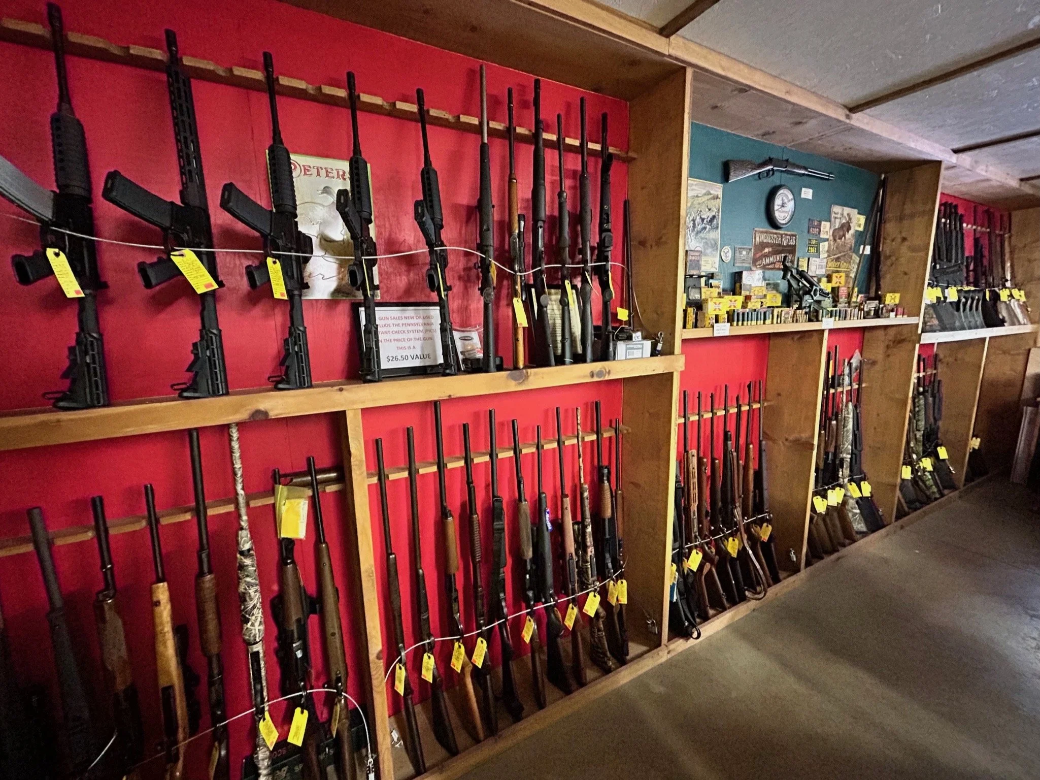 Multiple rifles and shotguns displayed on wooden shelves in a gun shop, with some tagged with yellow price tags and a sign indicating a value of $26.50.