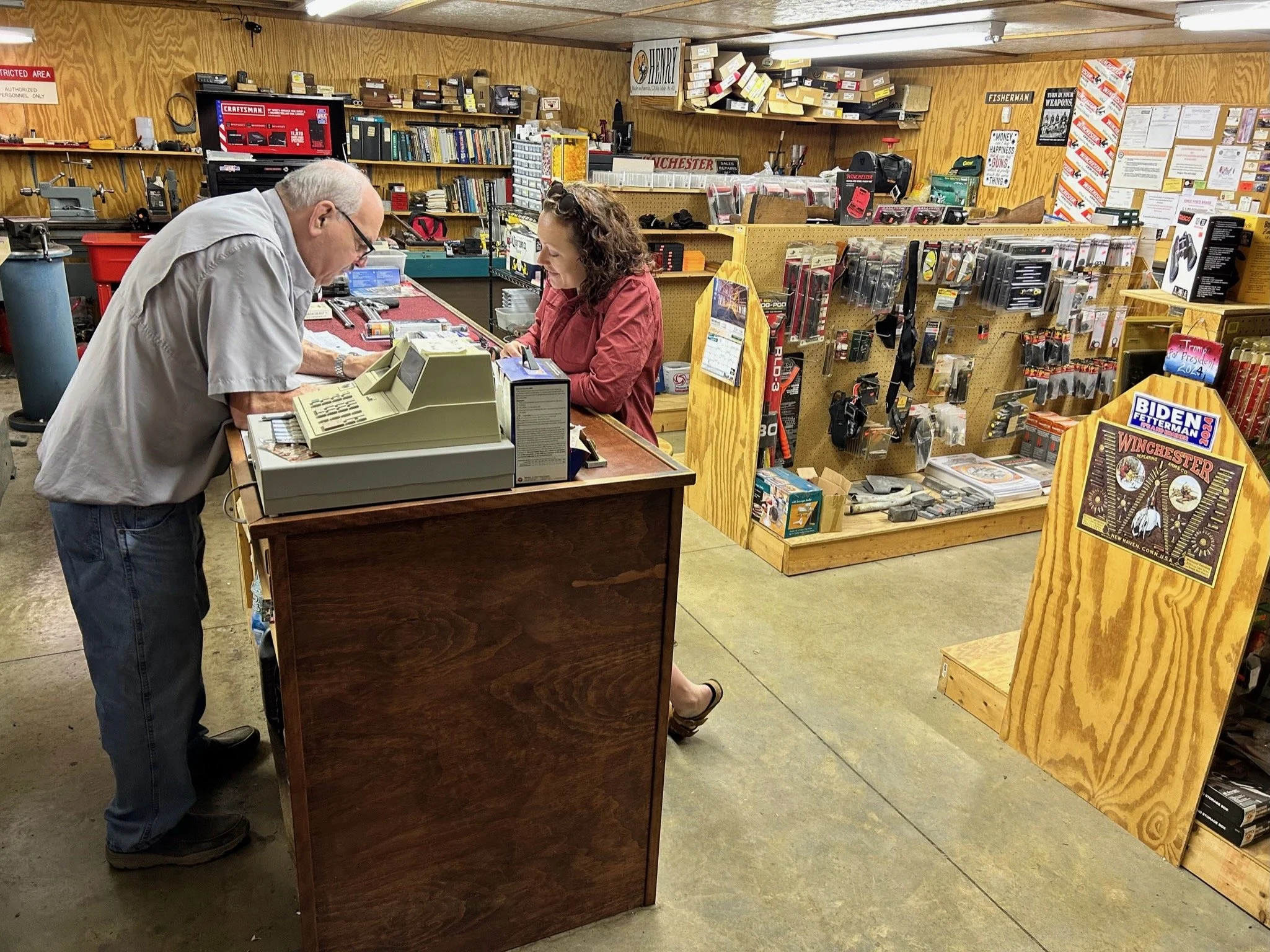 A man and woman standing at a store counter engaging in a transaction inside a hardware or sporting goods store, with shelves filled with tools, supplies, and promotional posters in the background.