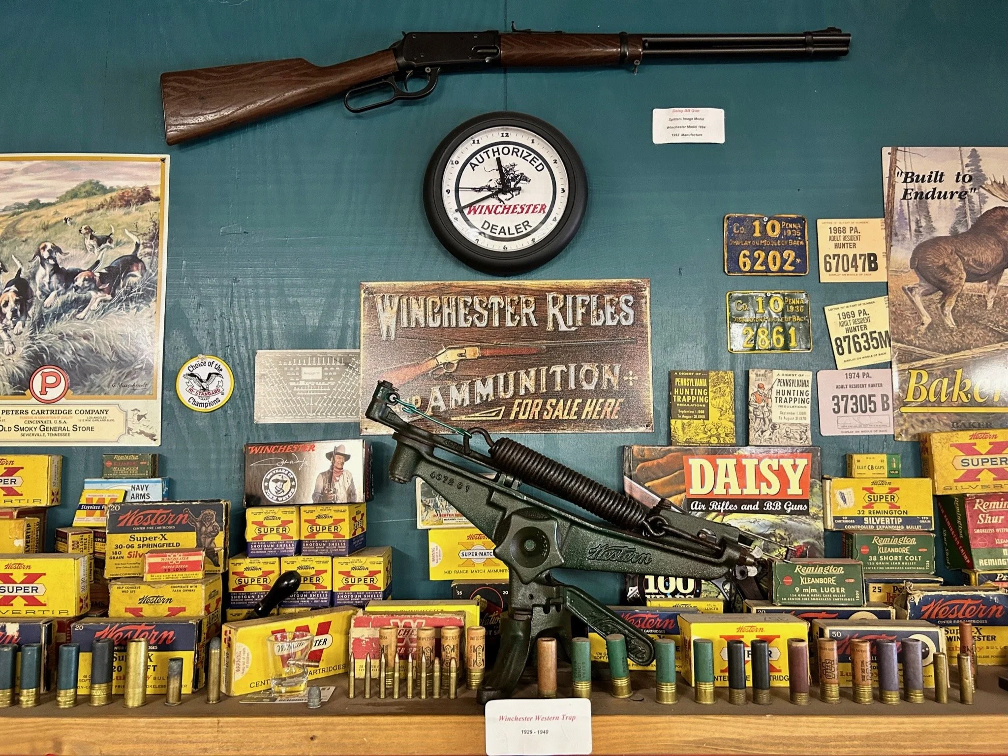 Display of Winchester firearms and ammunition, including a mounted rifle, boxed bullets, and various gun-related posters and signs.