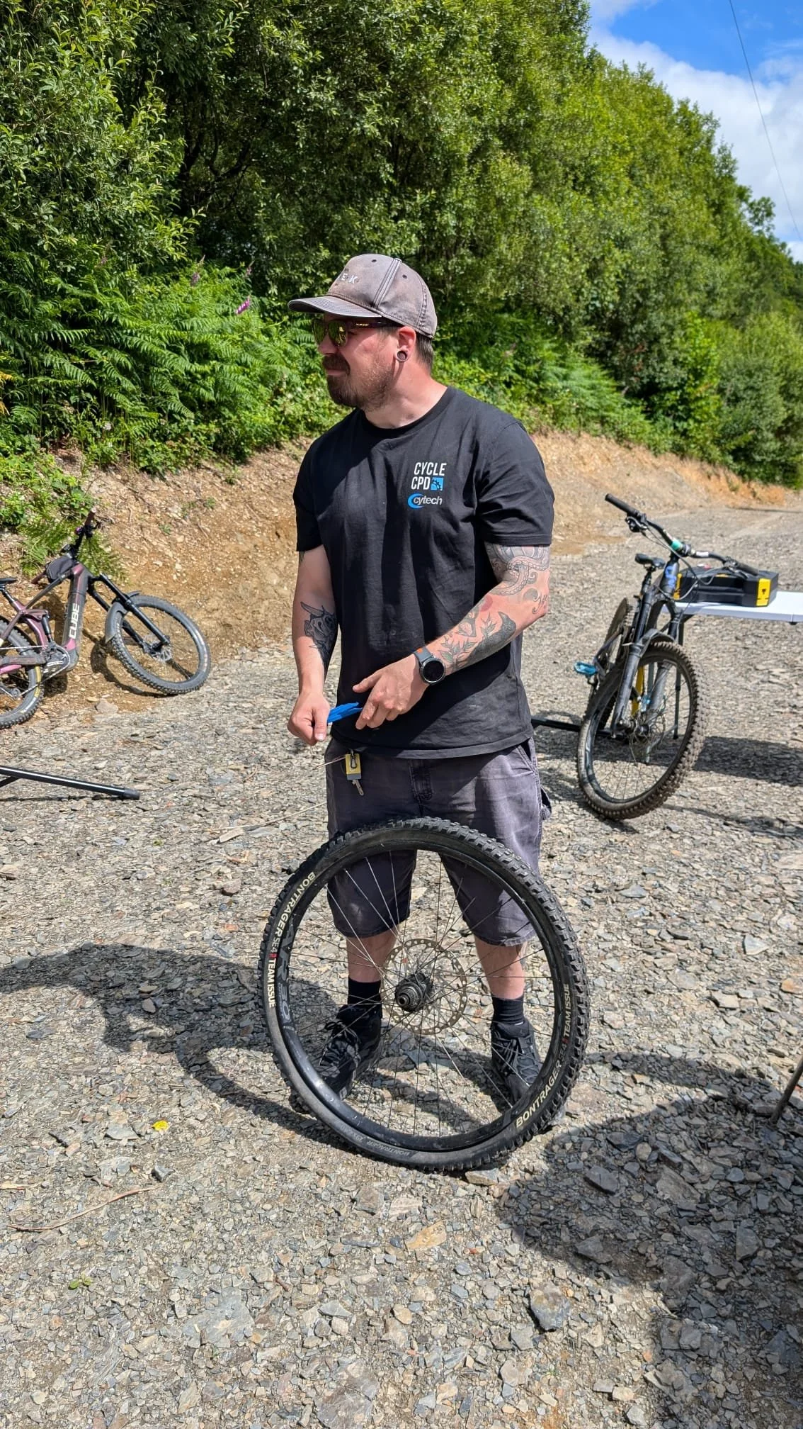A man wearing a gray cap, sunglasses, a black t-shirt, and gray shorts, standing outdoors on a gravel path, holding a bicycle wheel, with two other bicycles and green trees in the background.