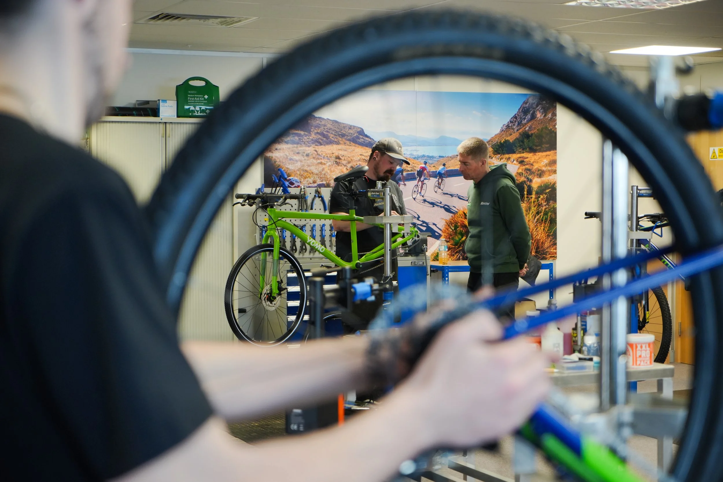 A bike shop with two men working on a green bicycle, seen through a foreground of several bicycle tires.