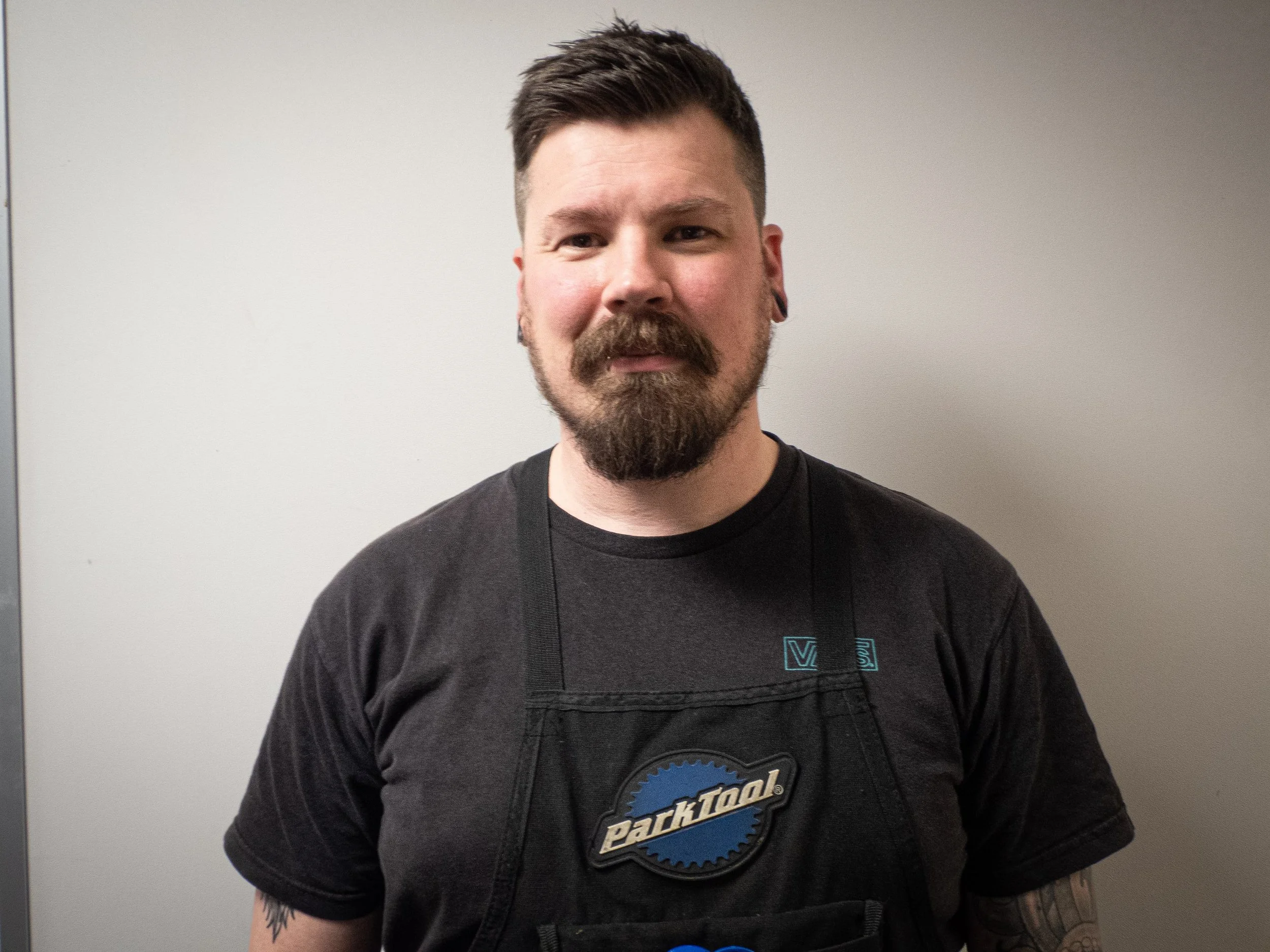 A man with short dark hair, a beard, and a mustache wearing a black T-shirt with a logo, standing against a plain white wall.