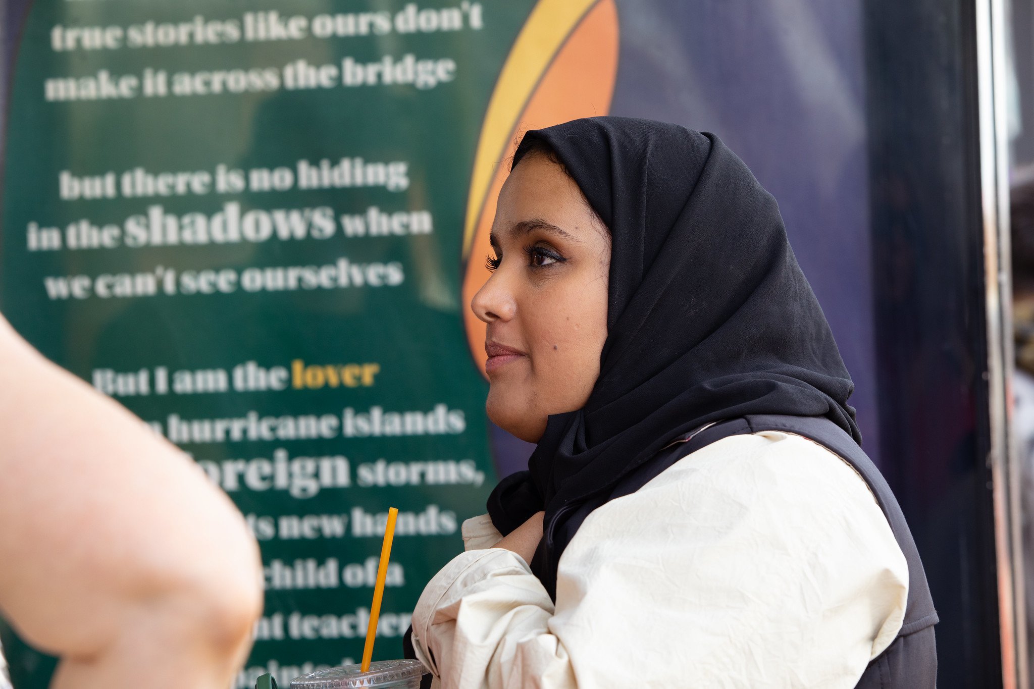 A person in a black headscarf and a white jacket, stands in front of a large bus stop poster board displaying poetry, with lines of its white text blurred but visible in the dark green background. They converse with someone offscreen to the left.