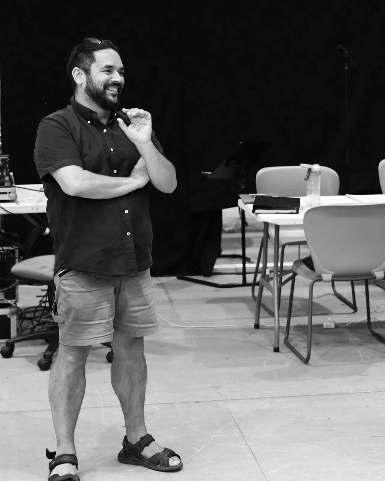 Black and white rehearsal image showing Keith smiling with his arms folded in a studio space with chairs and equipment in the background. Keith wears a buttoned shirt, shorts and sandals.