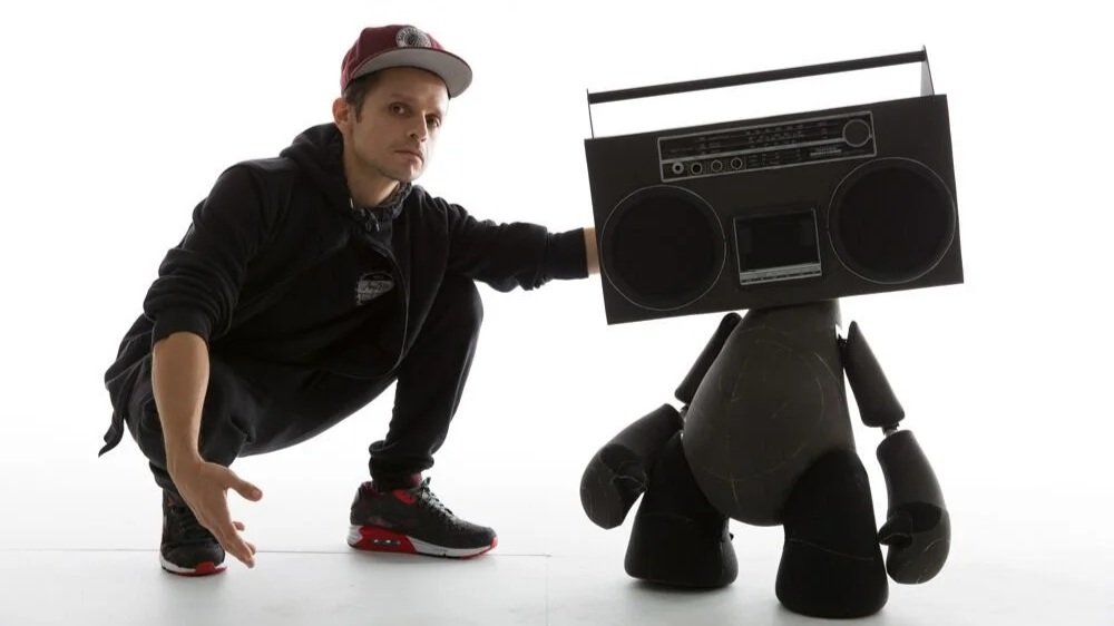 Promotional image showing a man crouched next to a large character-like sculpture with a boombox for a head, photographed against a white studio background. The man wears a red cap, a black hoodie and joggers and a red and black trainers.