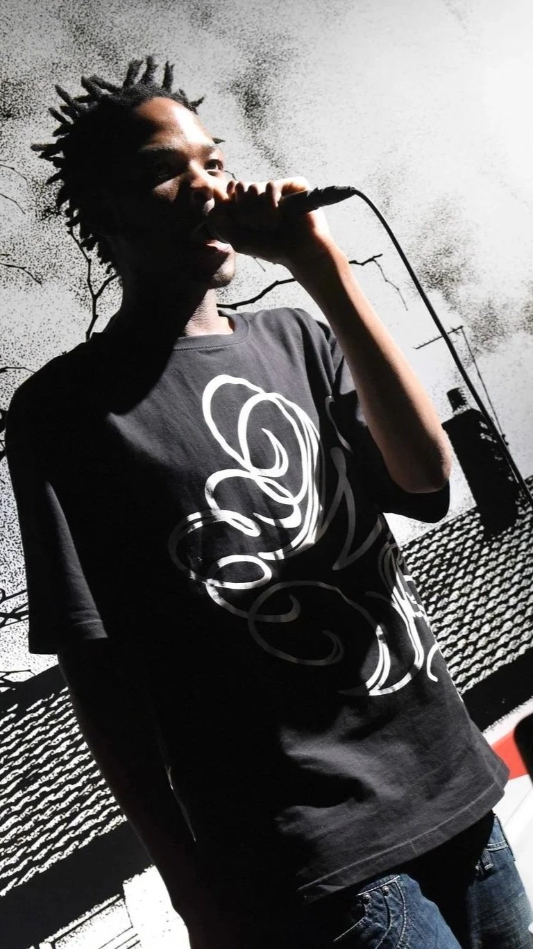 Production still of a performer vocalising into a handheld microphone, dressed in a dark T-shirt with a white swirling graphic on it. The performer is stood in front of a highly contrasted and black and white film grained image of a rooftop and cloud