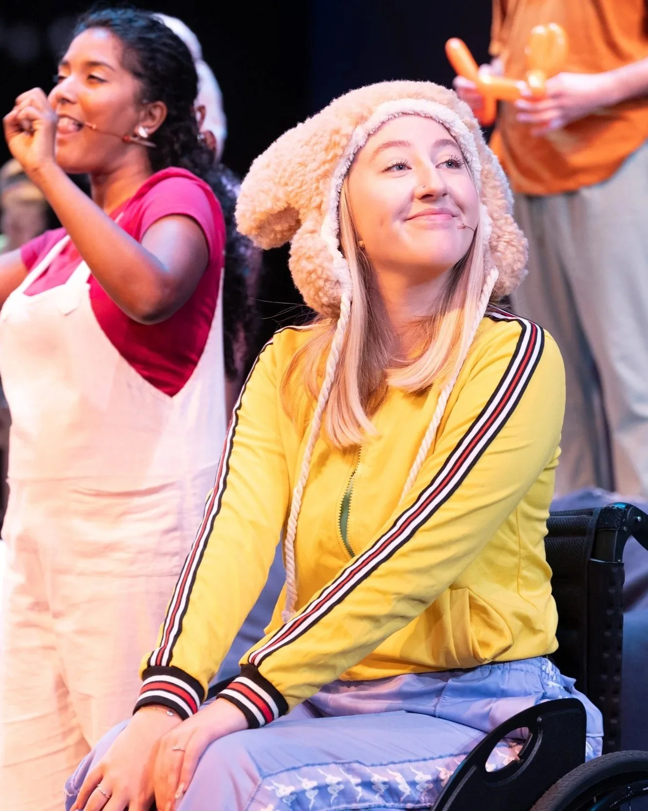 Production still of two performers on stage. A woman in a yellow top and fluffy bear hat sits in a wheelchair smiling in the foreground while another performer sings into a headset microphone behind her.