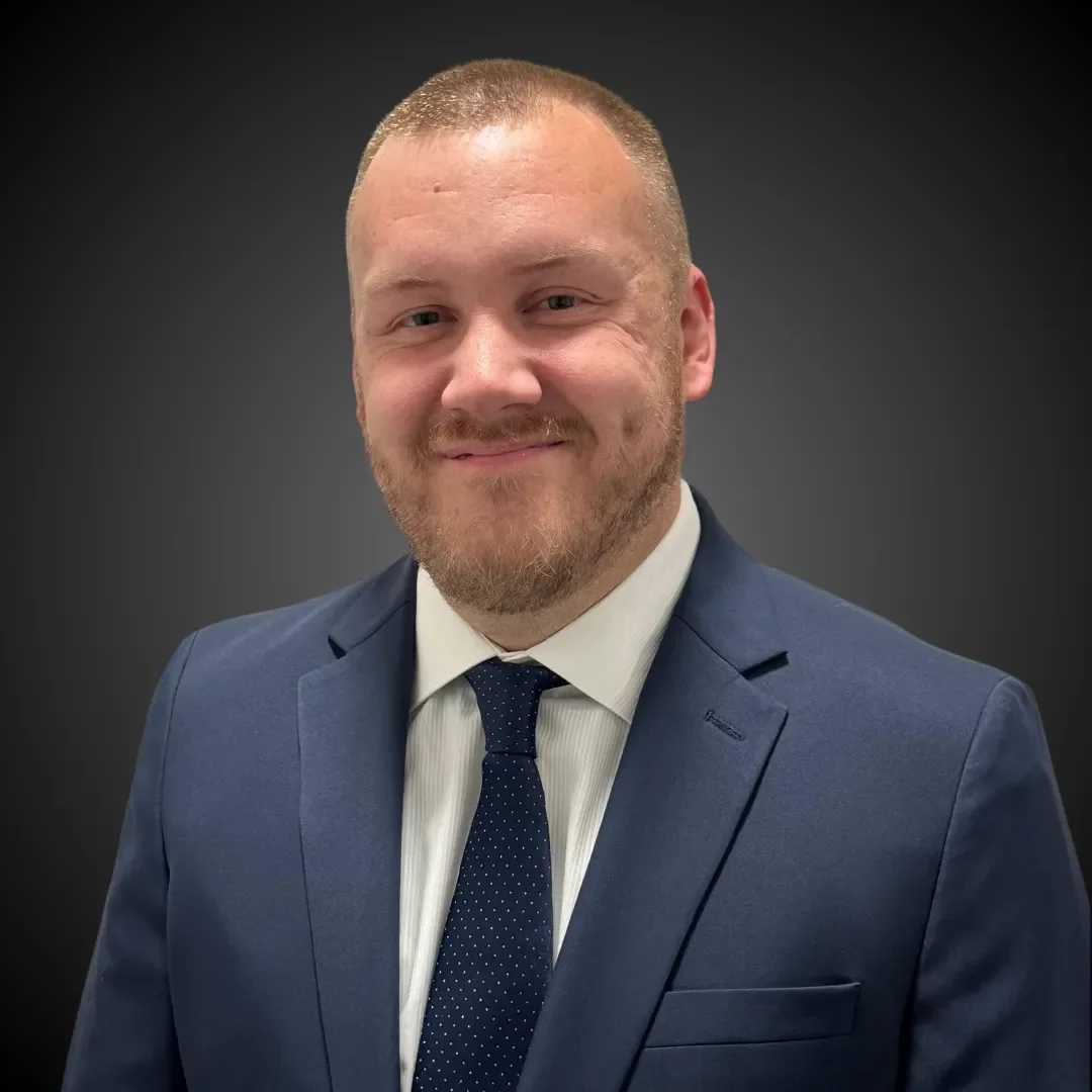 Headshot of a smiling man with light skin, short hair, and a beard, dressed in a blue suit and tie, against a dark gray background.