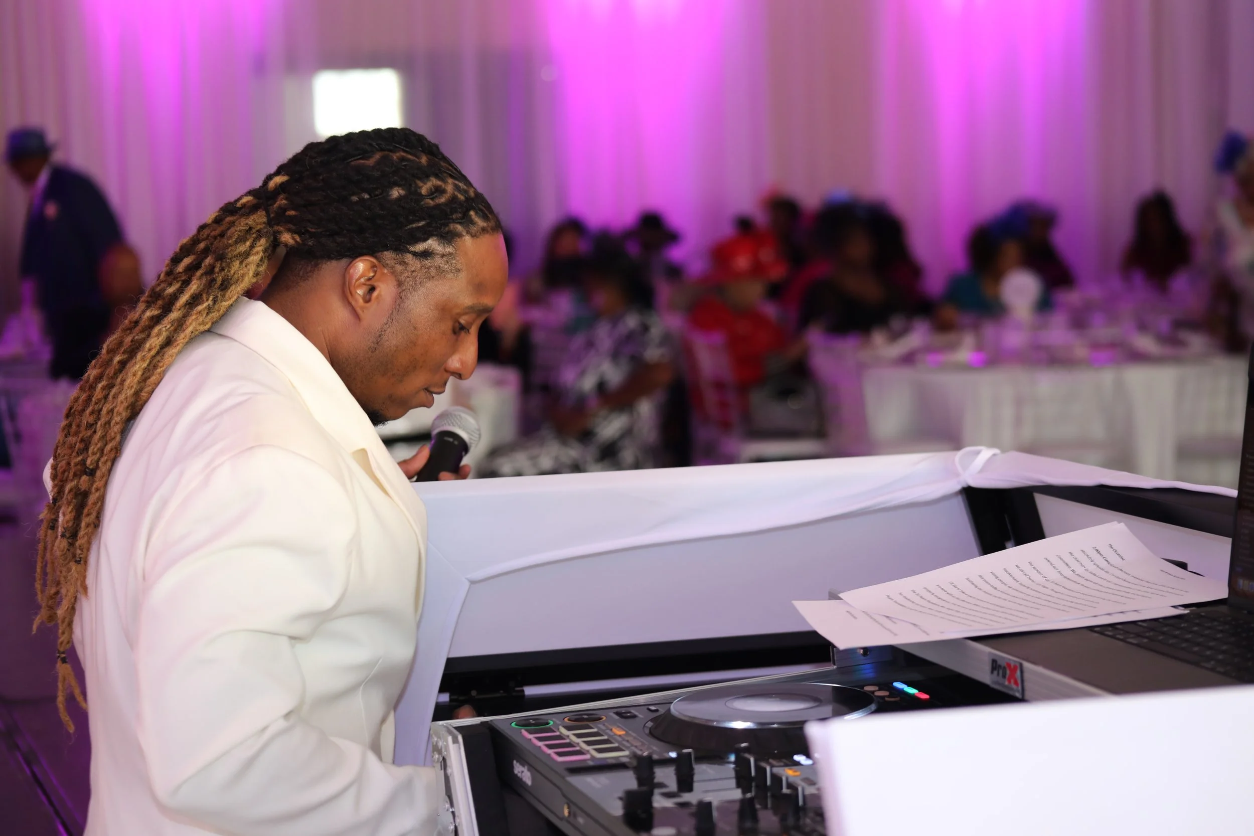 A DJ with long braids and a white suit is holding a microphone and standing behind a DJ console at a party or event, with a blurred audience in the background and festive lighting.