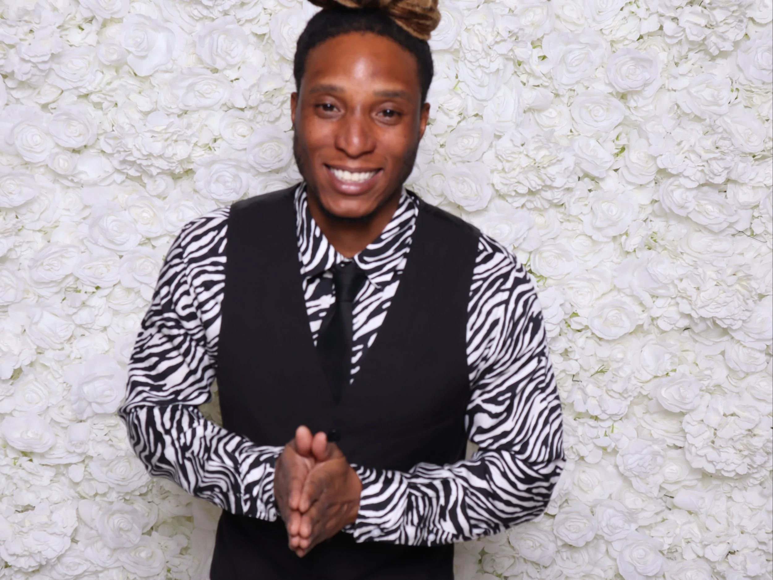 A smiling person with short hair and a beard, wearing a zebra print shirt, a black vest, and a black tie, standing in front of a white flower wall.
