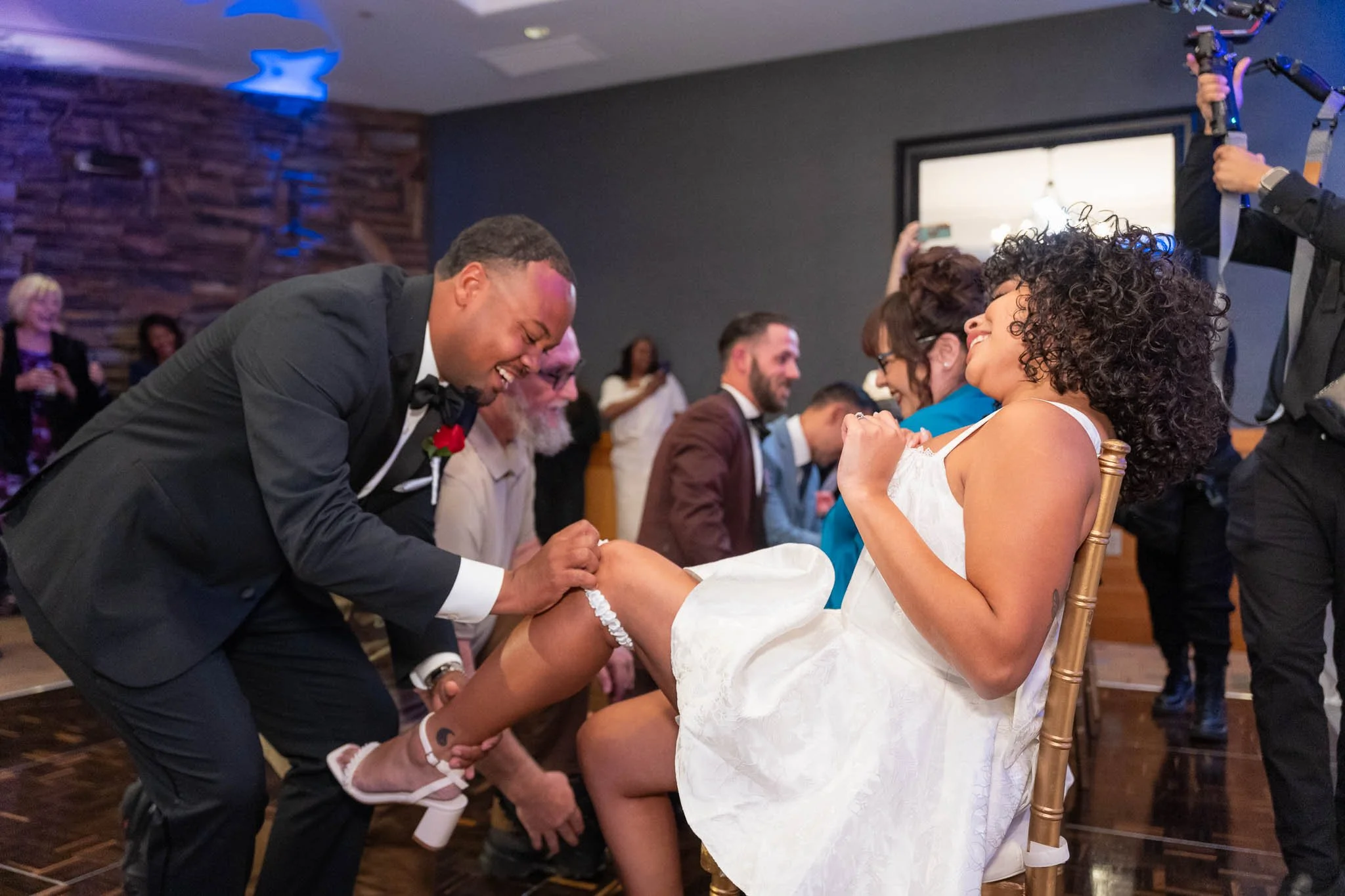 A man in a tuxedo is kneeling and laughing while pulling up a woman's white dress, revealing her leg and garter, during a wedding celebration. The woman is seated and smiling. Several other guests are watching and smiling in the background.