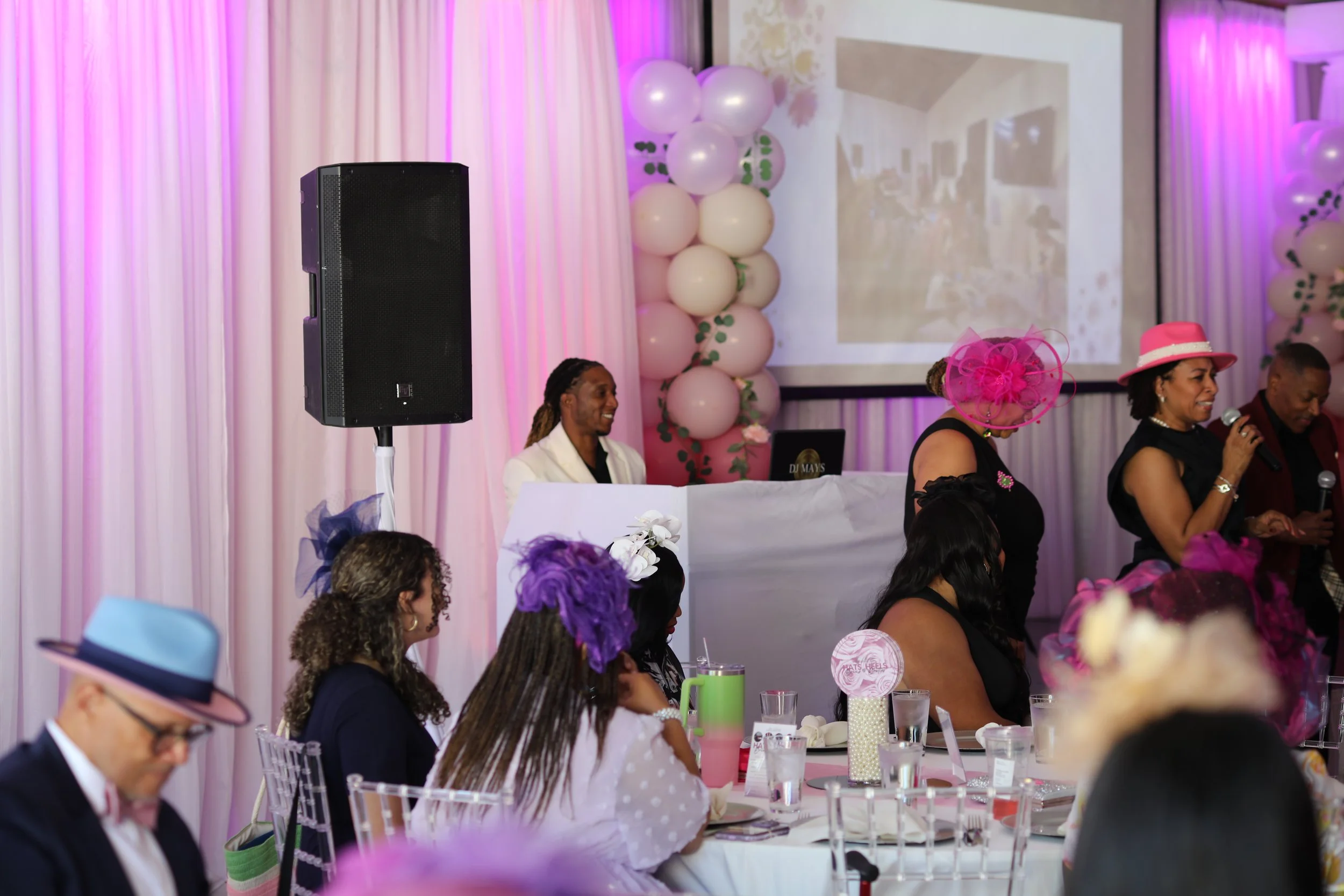 Guests seated at a decorated event space with pink and purple lighting, balloons, and a DJ setup, while a woman in a pink hat speaks into a microphone.