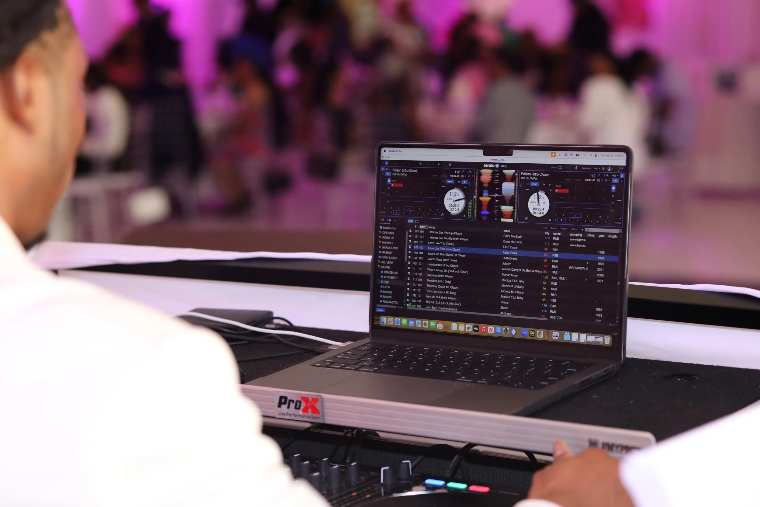 A DJ or music performer at a party with a laptop displaying DJ software, a mixer, and a crowd dancing in the background with pink and purple lighting.
