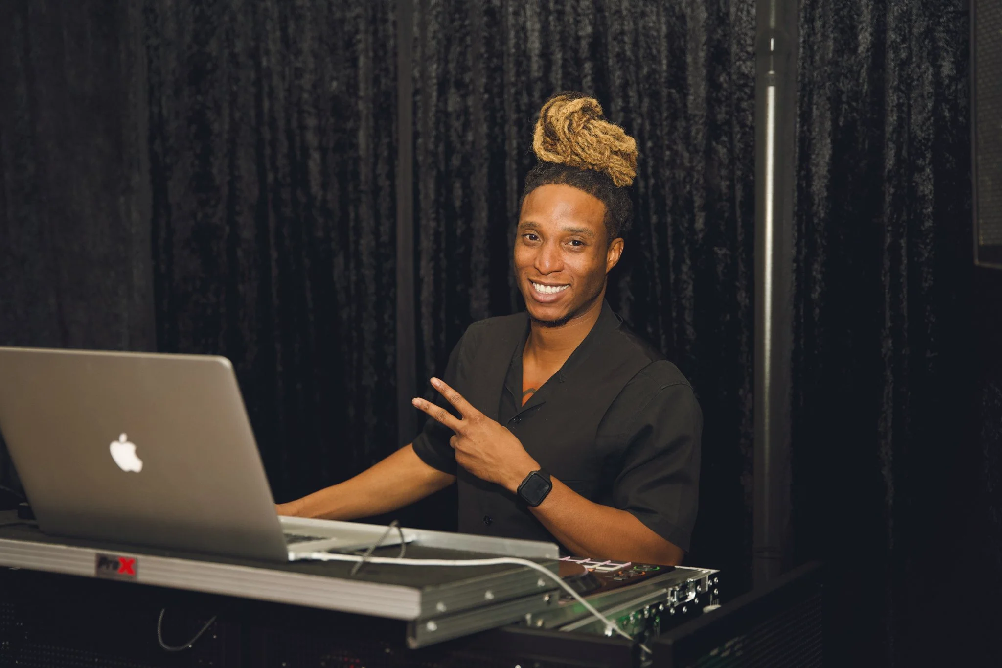A smiling DJ with blonde dreadlocks tied up, making a peace sign with his right hand while sitting at a DJ booth with a laptop.