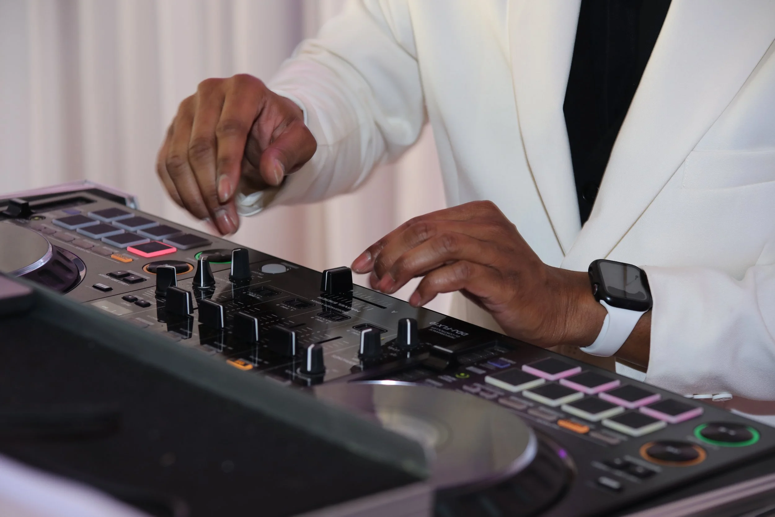 Close-up of a DJ's hands operating a professional DJ controller with various knobs, buttons, and a turntable, wearing a white blazer and a smartwatch.