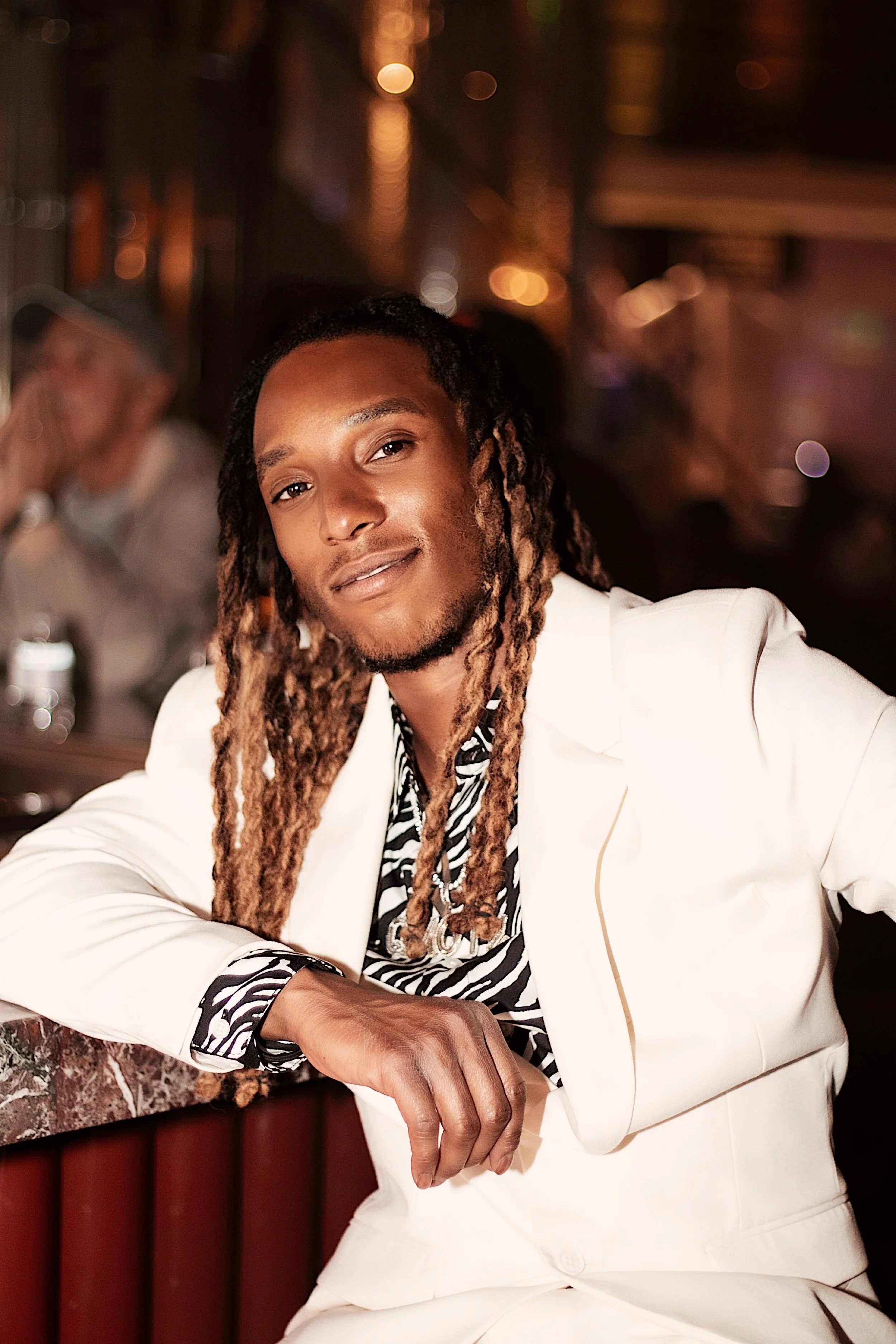 A man with long dreadlocks and a goatee, dressed in a white suit and a zebra-patterned shirt, sitting at a bar in a dimly lit venue with warm lighting and blurred background of other patrons.
