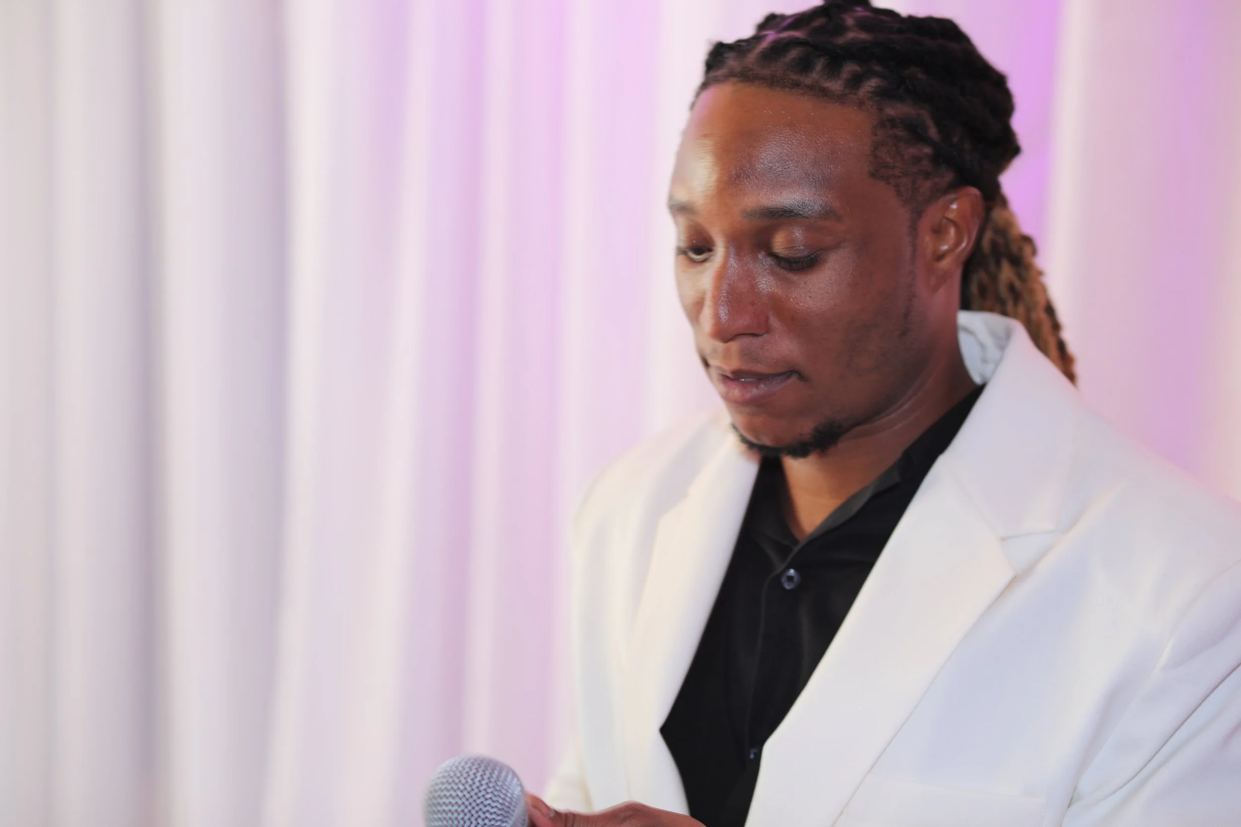 Man in a white suit holding a microphone, looking down, with a light-colored curtain background.