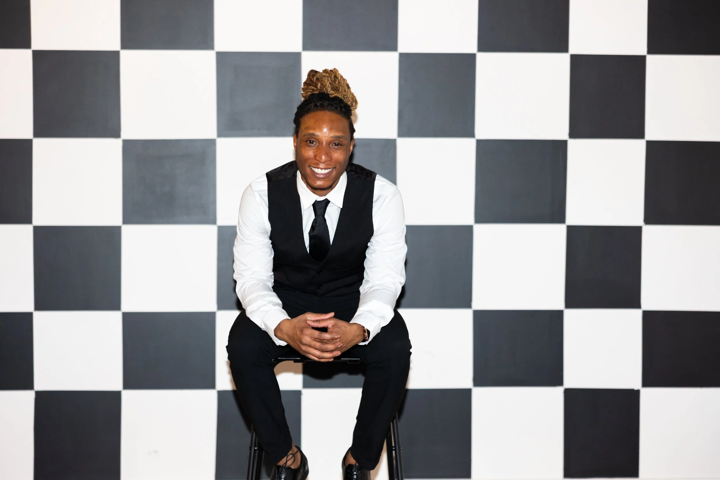 A smiling man dressed in a white shirt, black vest, black tie, and black pants sitting on a chair in front of a black-and-white checkered wall.