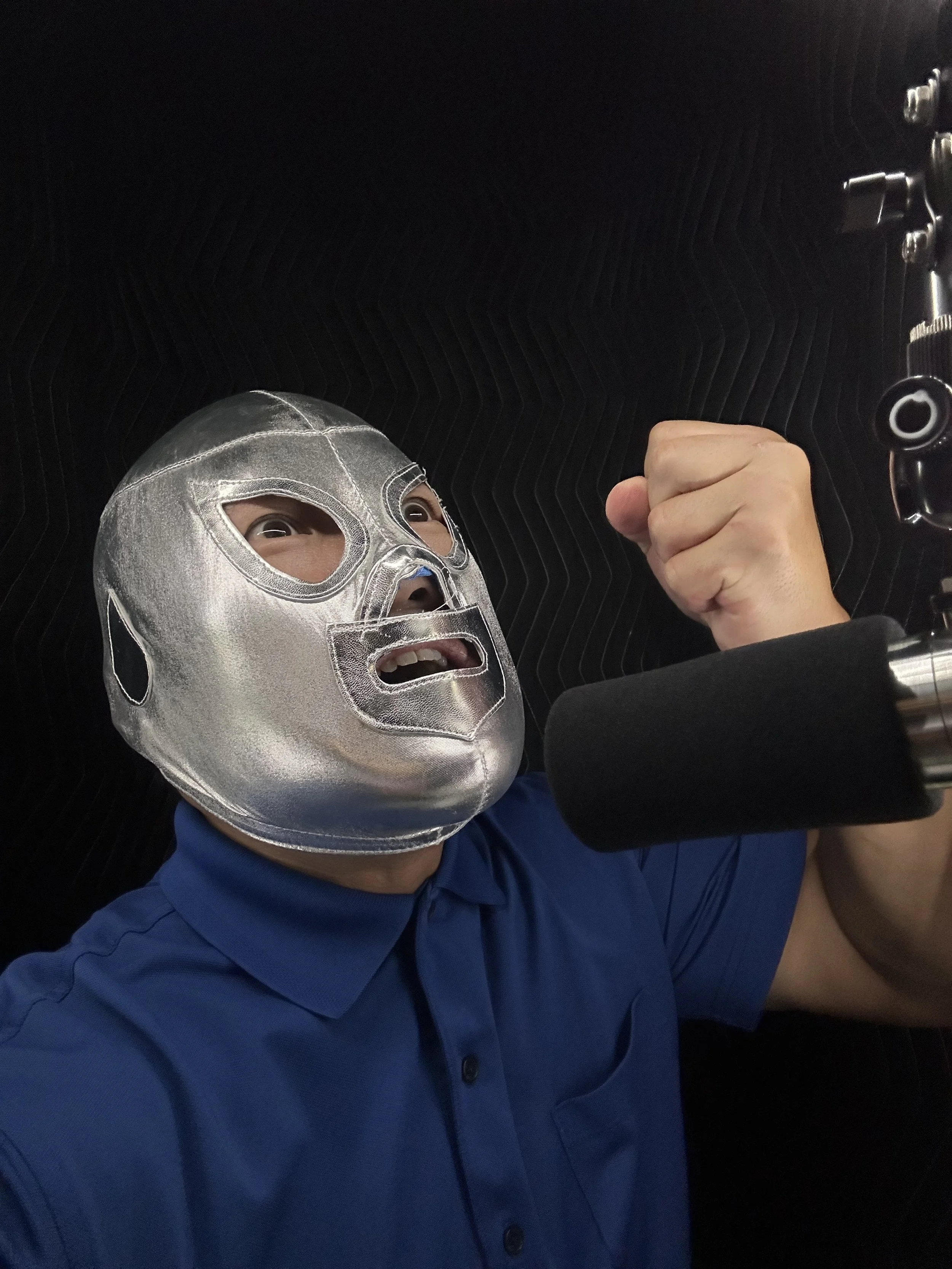 Person wearing a silver luchador mask, blue shirt, and fist raised, speaking into a microphone.