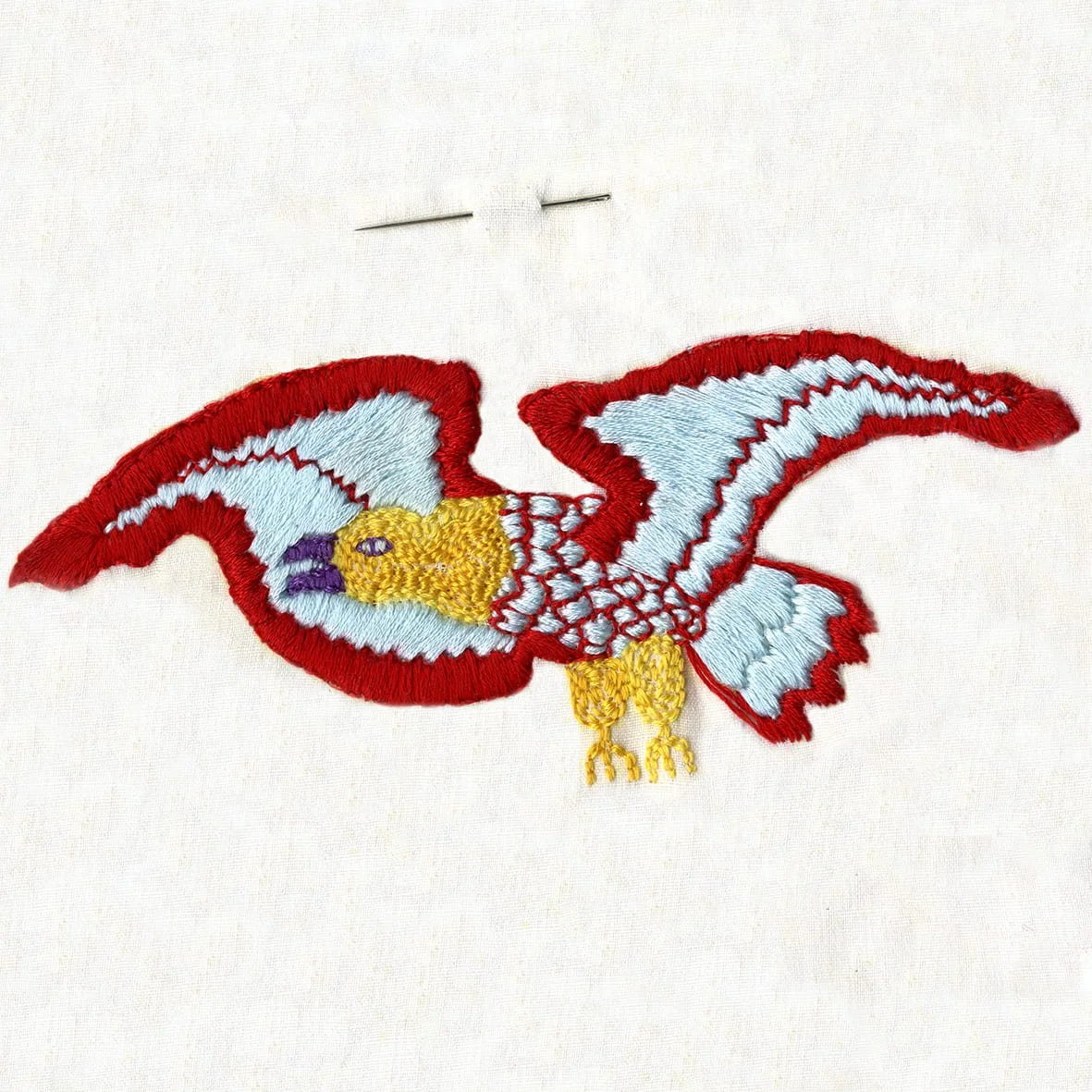 Broderie d'un oiseau avec des ailes déployées, principalement en bleu, rouge, jaune et violet, sur un fond blanc.