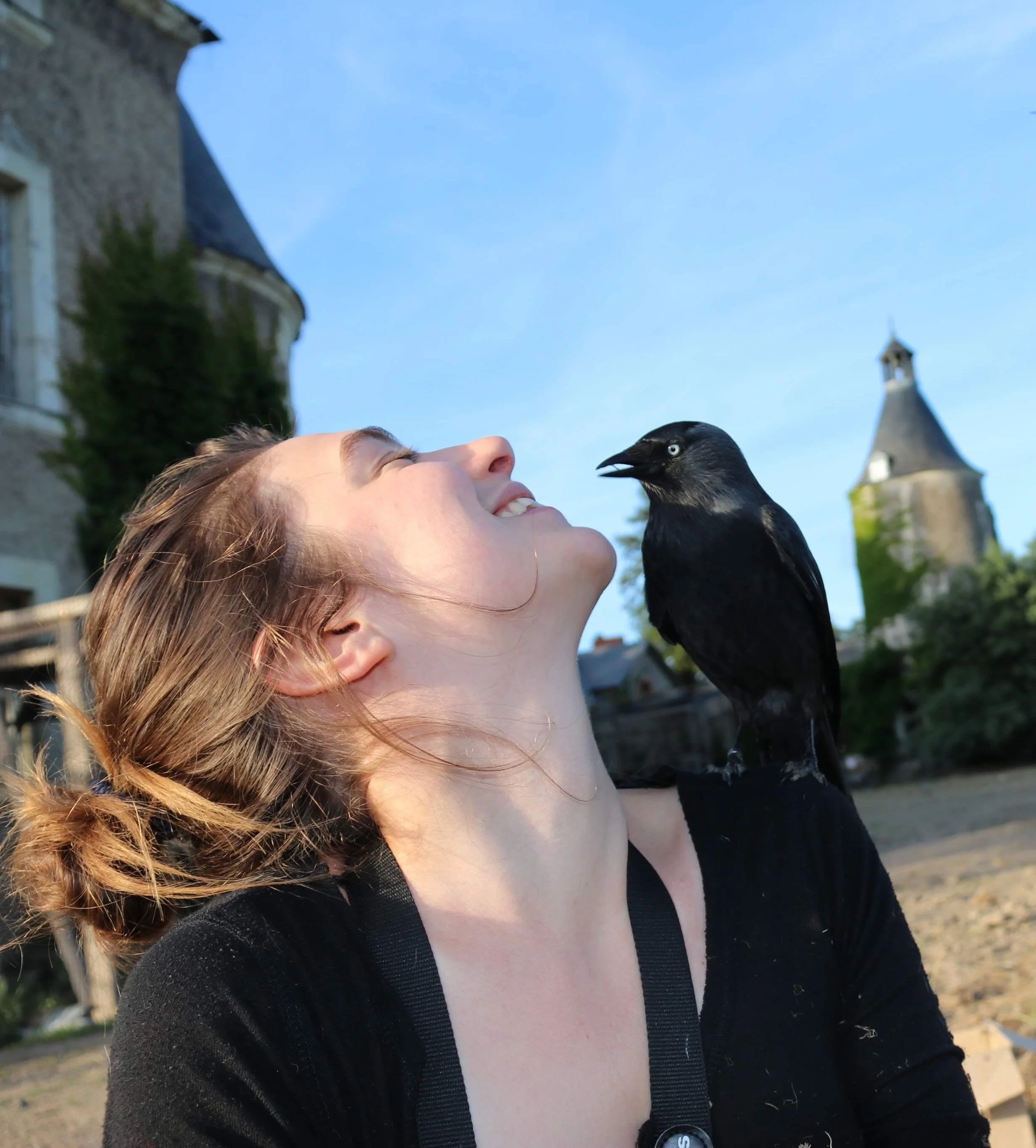 Une femme souriante avec un oiseau noir perché sur son épaule, en extérieur, avec un ciel bleu et un bâtiment ancien en arrière-plan.