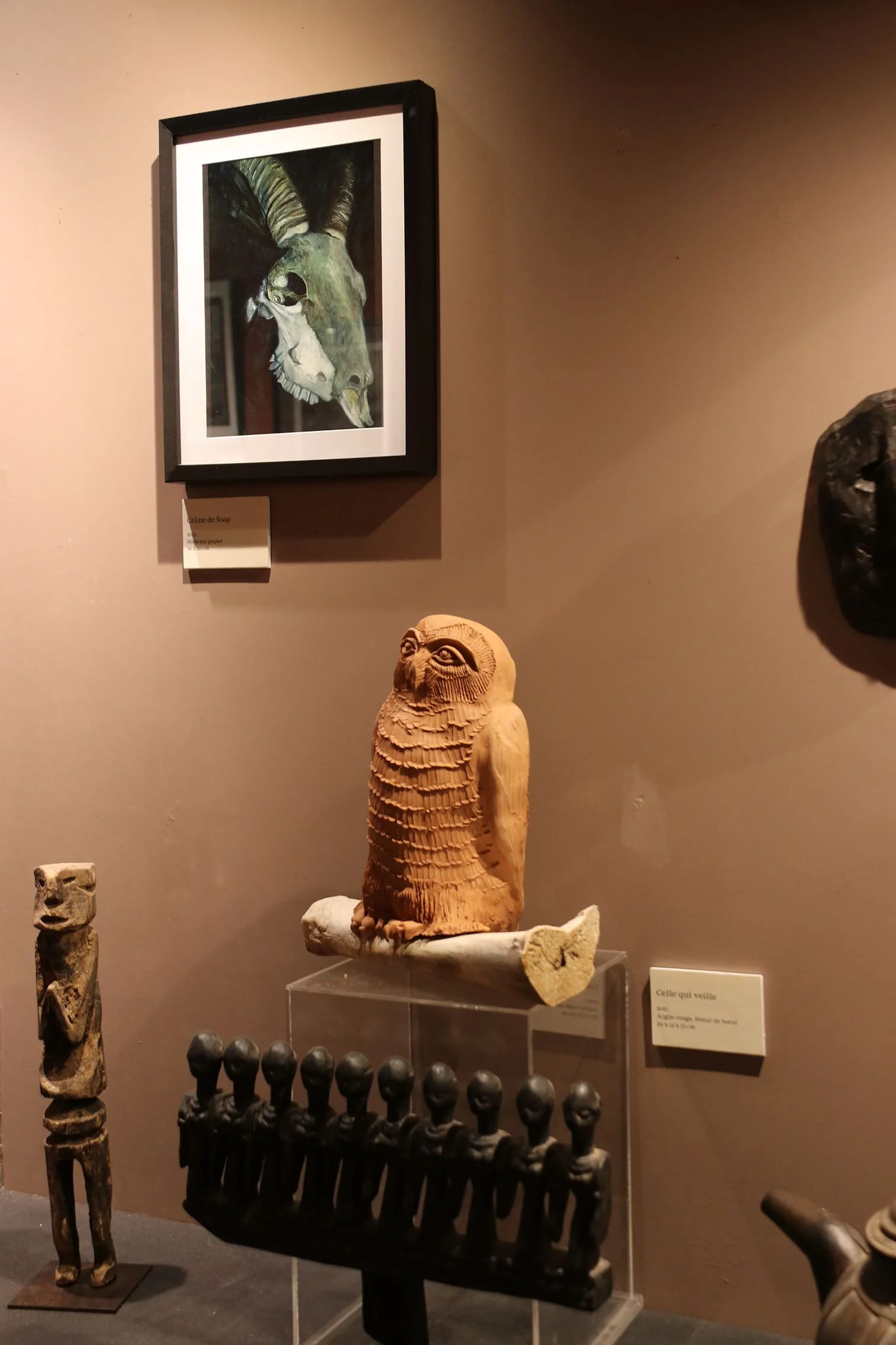 Collection d'objets artistiques et sculpturaux dans un musée, comprenant une peinture encadrée d'un animal hybride, une statue en bois représentant un hibou, et plusieurs figurines en pierre ou en bois.
