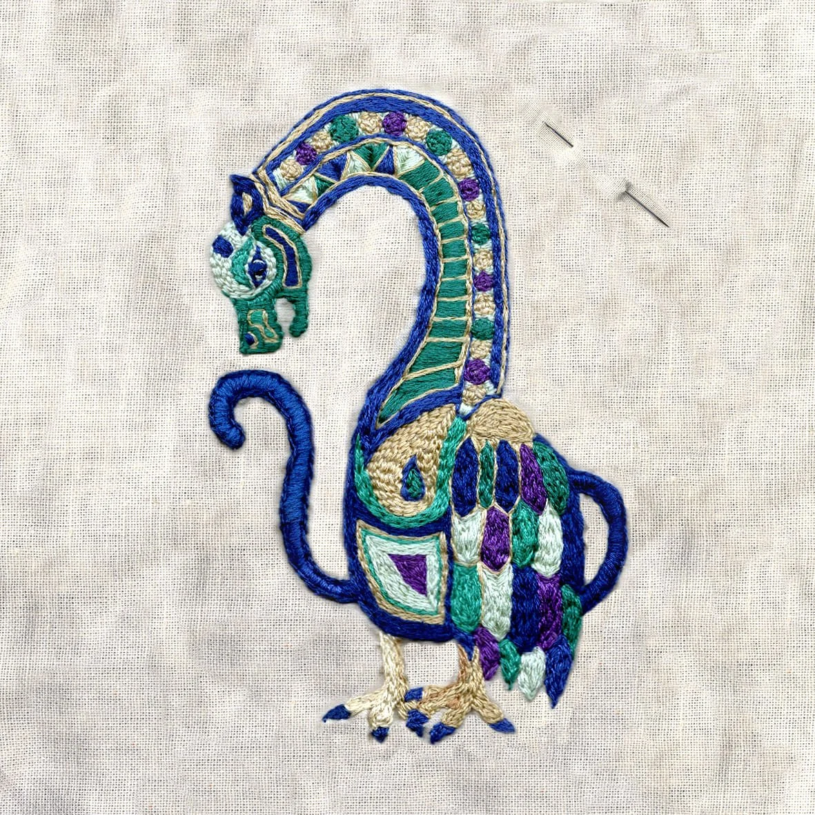 Broderie représentant un paon stylisé avec des couleurs vives comme le vert, le violet, le beige et le bleu sur un fond en tissu beige.