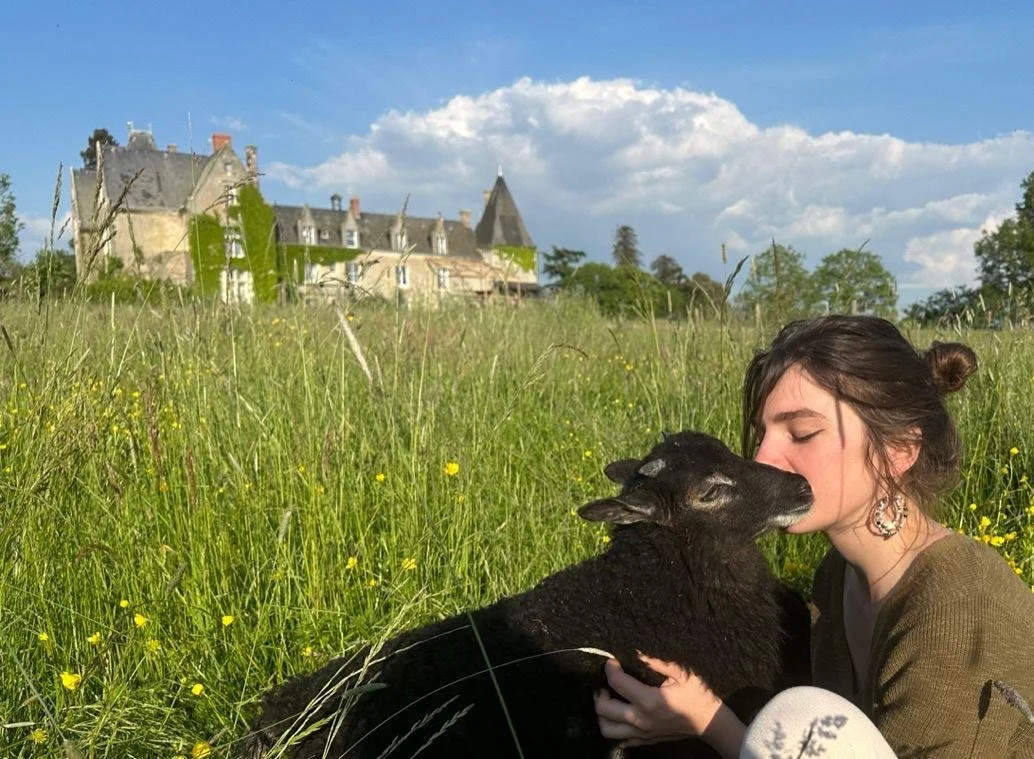 Une jeune femme embrasse une petite chèvre noire dans un champ vert avec un château en arrière-plan sous un ciel en partie nuageux.