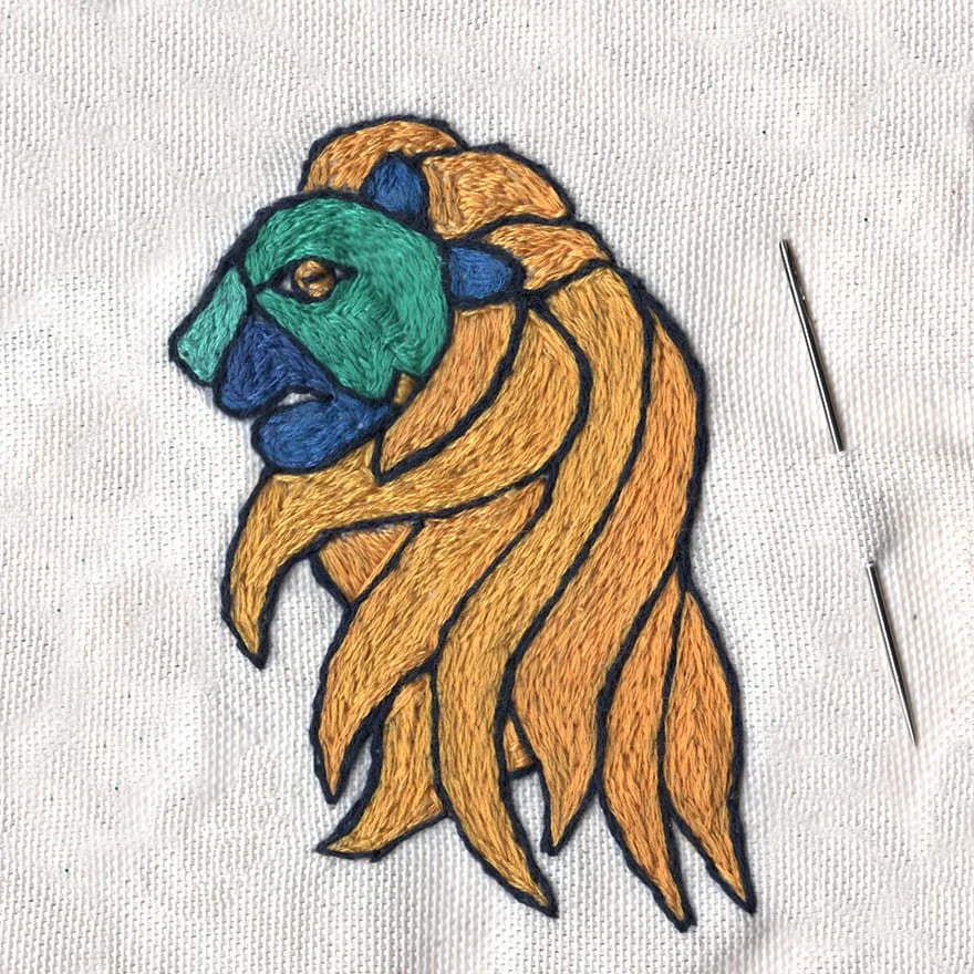 Broderie d'un lion stylisé avec des couleurs vives sur une toile blanche, avec deux aiguilles à coudre à côté.