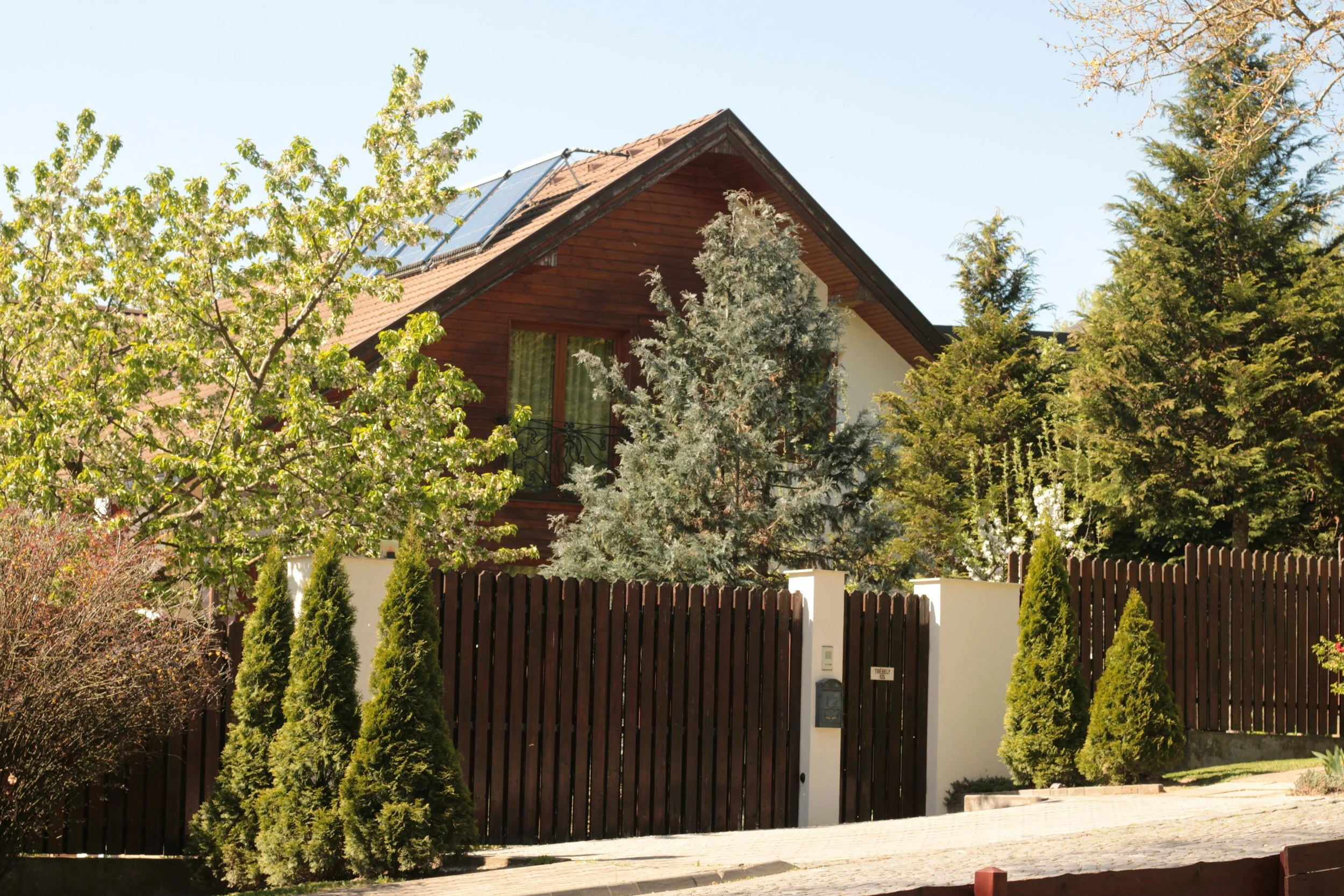 A house with a brown roof and wooden exterior, surrounded by trees and a dark wooden fence with a white pillar and gate.