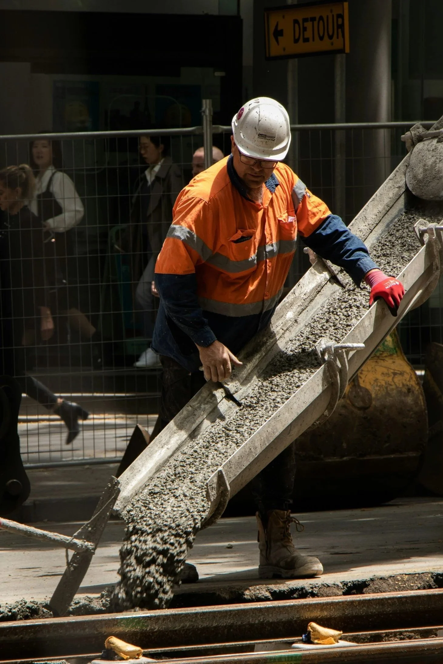 A construction worker in an orange safety jacket, white helmet, and red gloves is pouring concrete onto a railway track at a construction site, with a city background and pedestrians behind a safety barrier.
