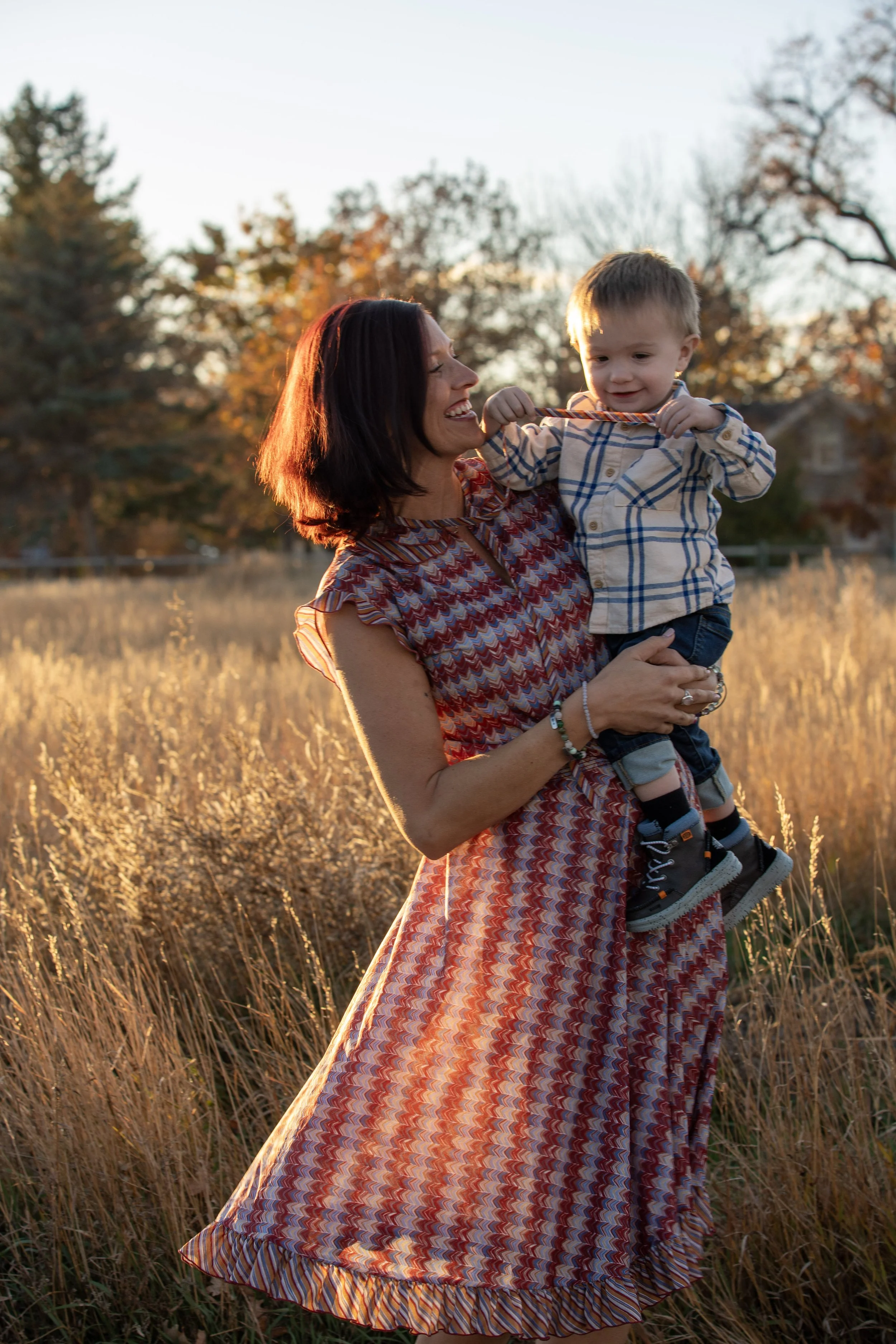 A woman holding a young boy in a grassy field during sunset, with trees and houses in the background.