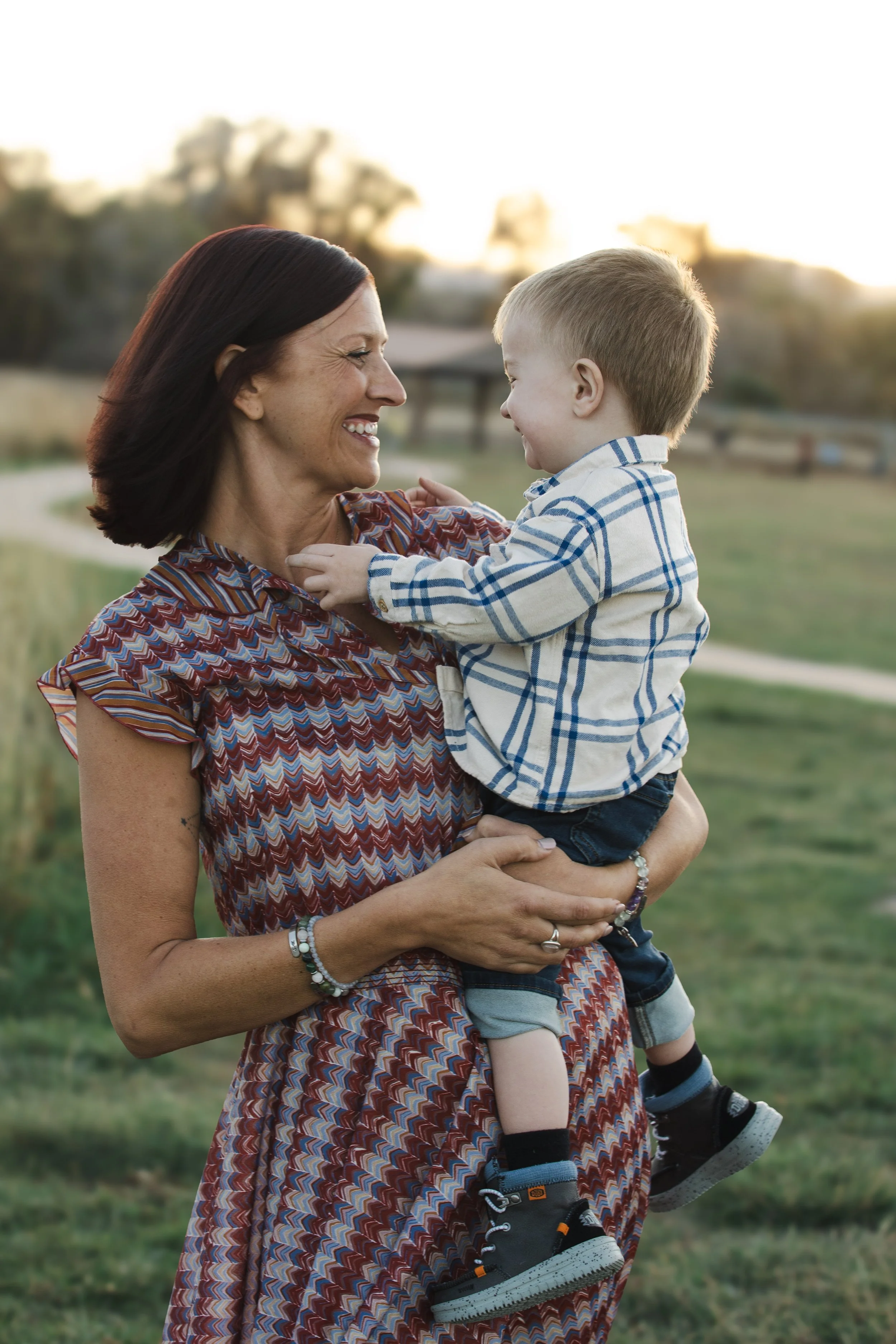 A woman holding a young boy outdoors during sunset, both smiling and looking at each other.