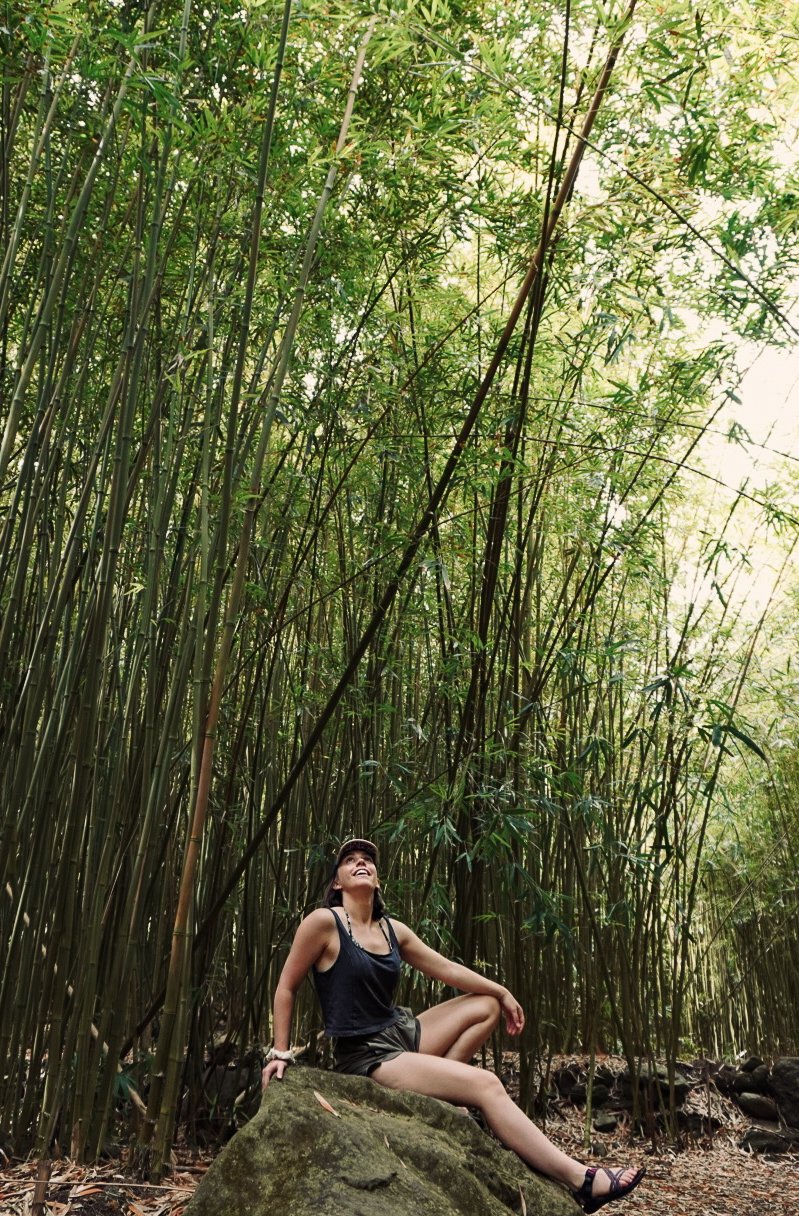A woman sitting on a large rock in a bamboo forest, smiling and looking up, surrounded by tall bamboo stalks and greenery.