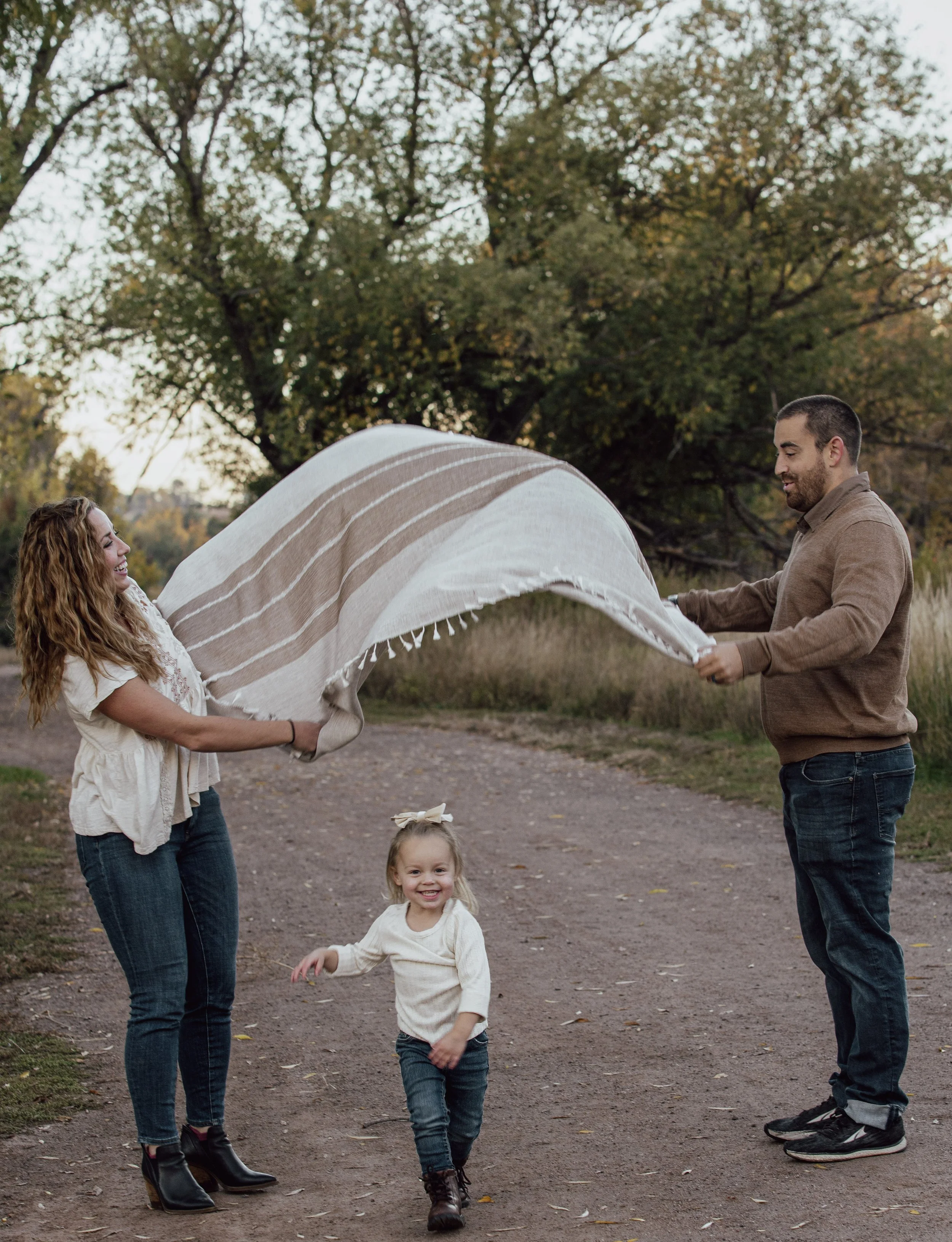 A family of three outdoors on a dirt path, with trees in the background. A woman and man are holding a blanket, creating a makeshift tent, while a young girl runs beneath it, smiling.