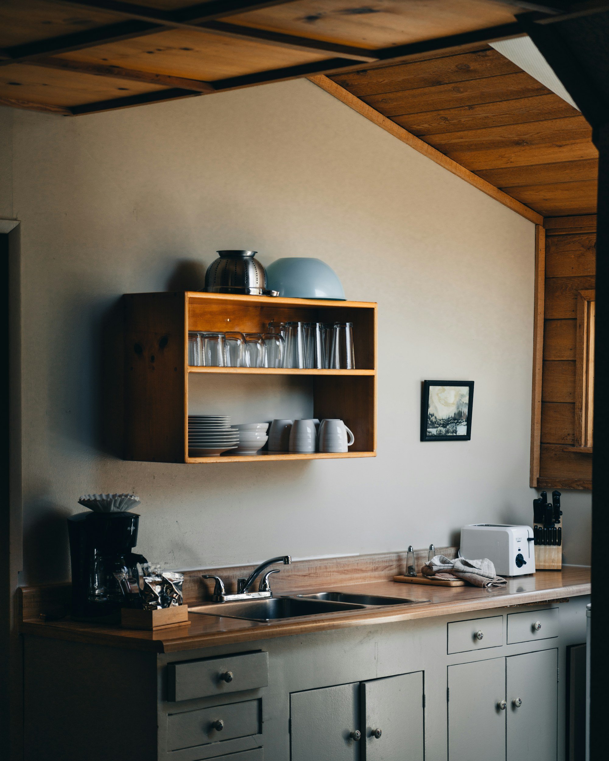 A kitchen with green cabinets, a wooden countertop, a double sink, and a wooden shelf holding glasses, cups, and a pot. There is a coffee maker, a toaster, and a knife block on the counter. A small framed picture hangs on the wall.