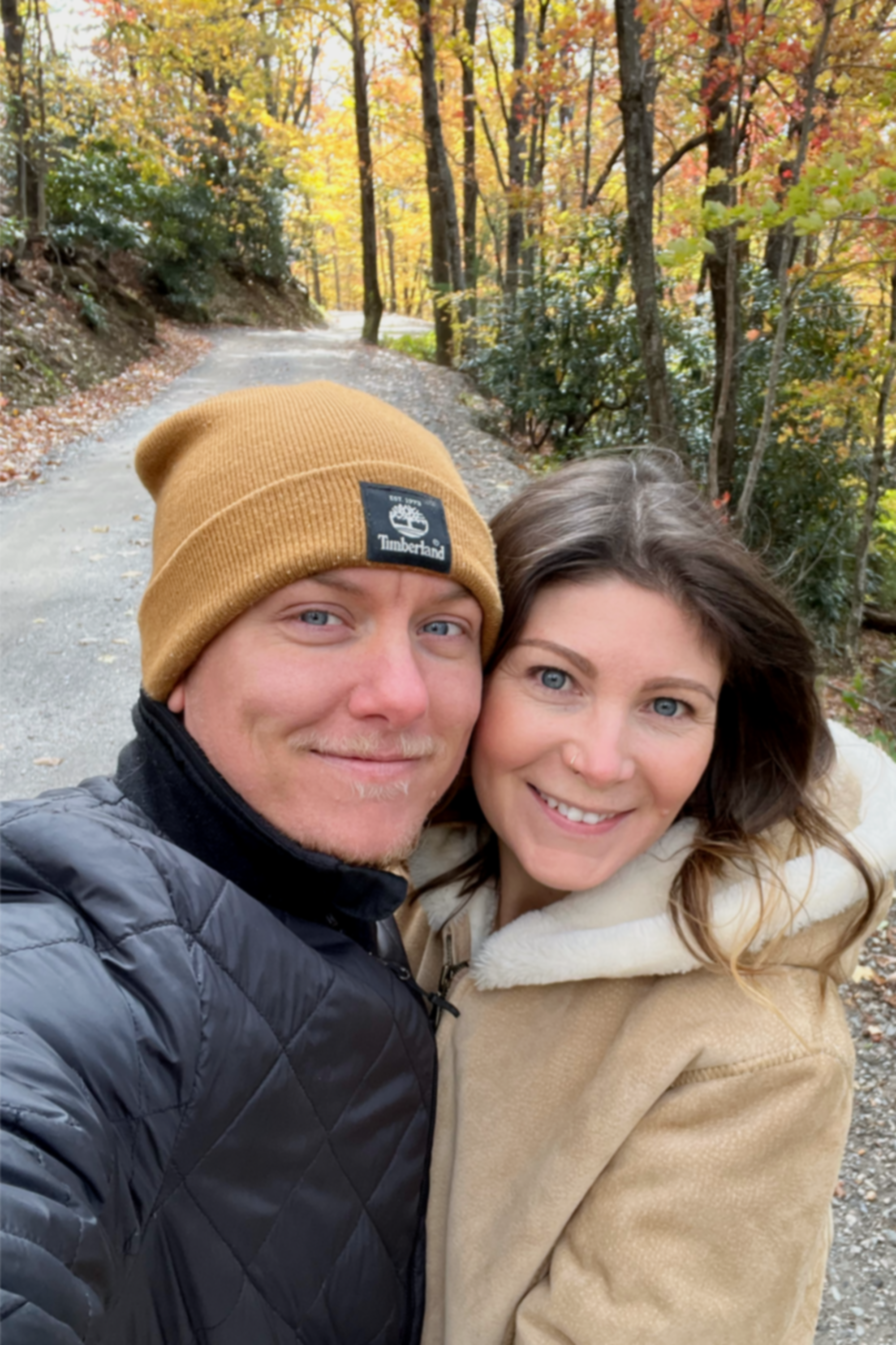 A smiling man and woman taking a selfie together outdoors on a forest trail with autumn-colored trees in the background.