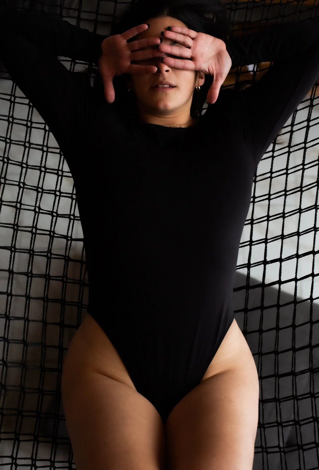 Une femme allongée sur un lit avec un drap noir, portant un body noir, les mains couvrant ses yeux.