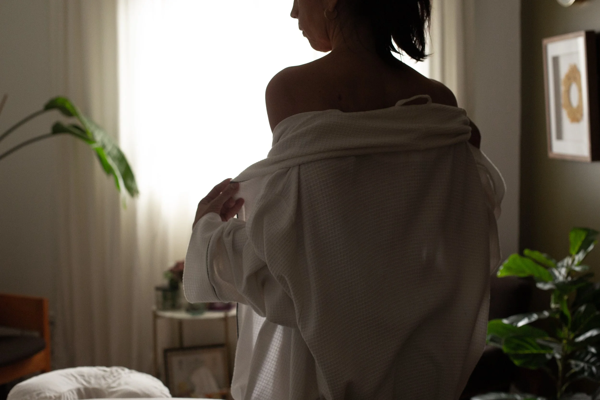 Une personne en sous-vêtements est en train de mettre un kimono blanc dans une pièce lumineuse avec des plantes et des cadres sur le mur.