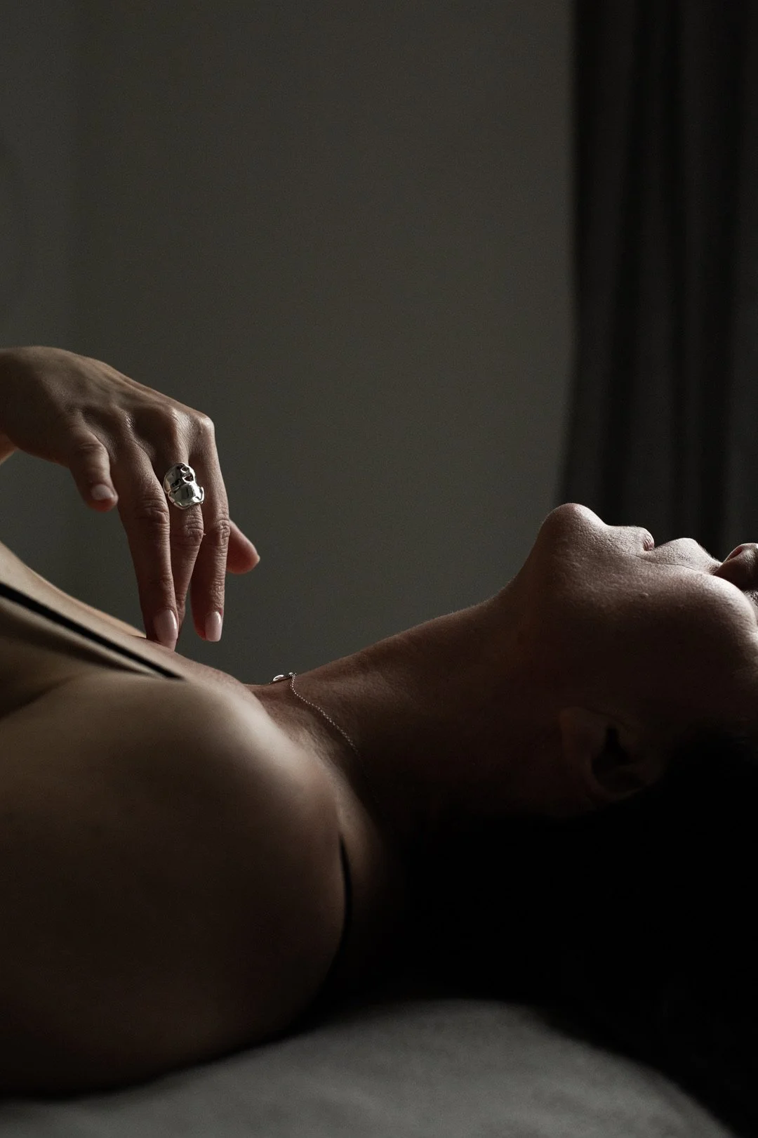 Une personne allongée sur un lit avec le visage tourné vers le haut, peau éclaircie, portant un collier fin et un bague en forme de crâne. La lumière est douce et sombre, avec un fond flou de rideaux noirs.