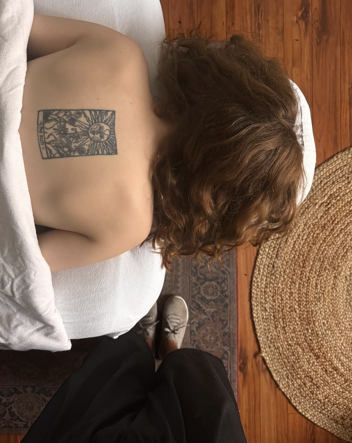 Une personne allongée sur un lit vu de dos, avec une courte mèche de cheveux bouclés rouges, portant un tatouage sur le dos représentant une illustration religieuse ou mythologique, avec un sol en bois et un tapis rond en rotin visible à côté.