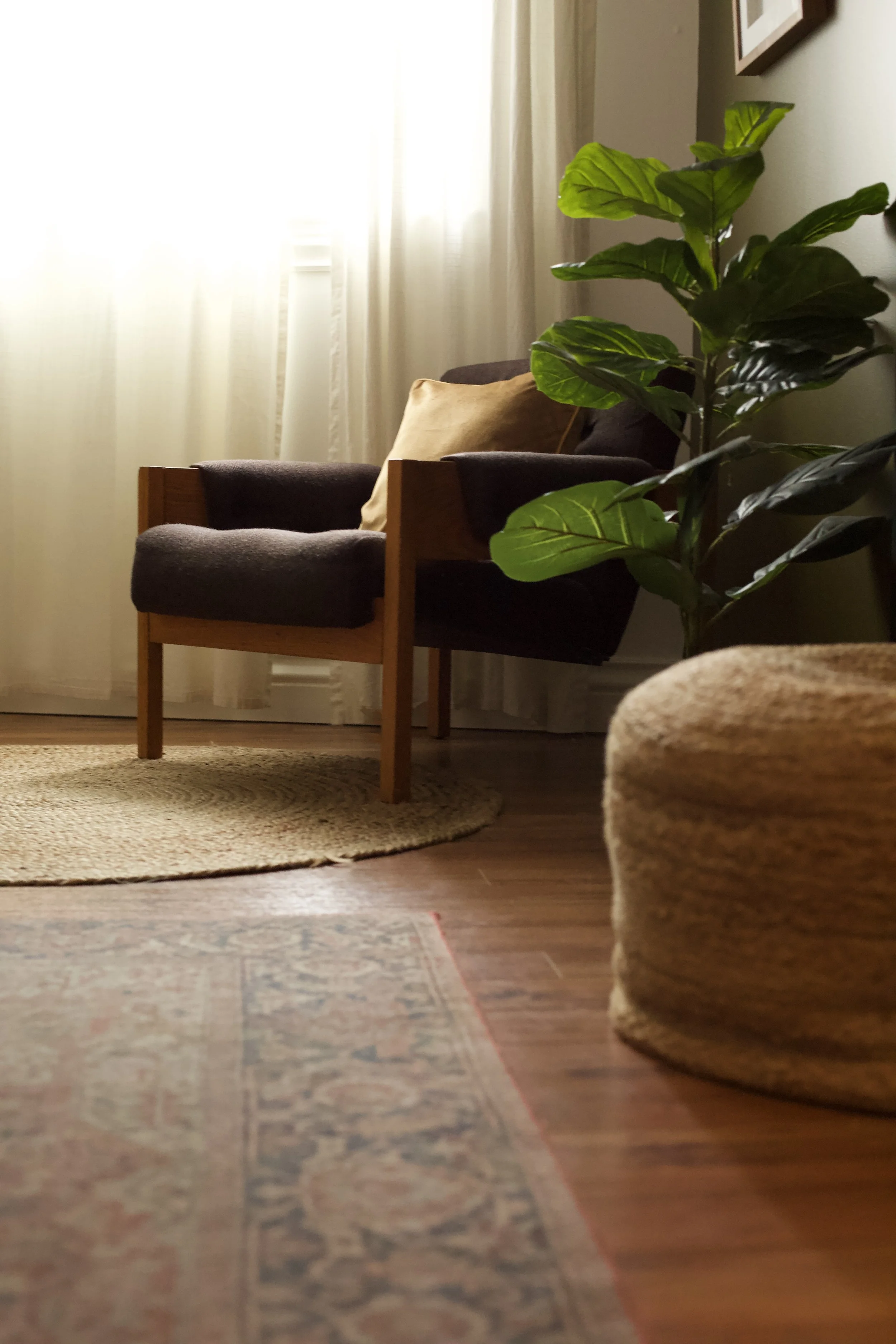 Coin salon avec fauteuil en bois, coussin beige, végétation verte, rideaux légers, tapis rond en jute et tapis traditionnel en laine.